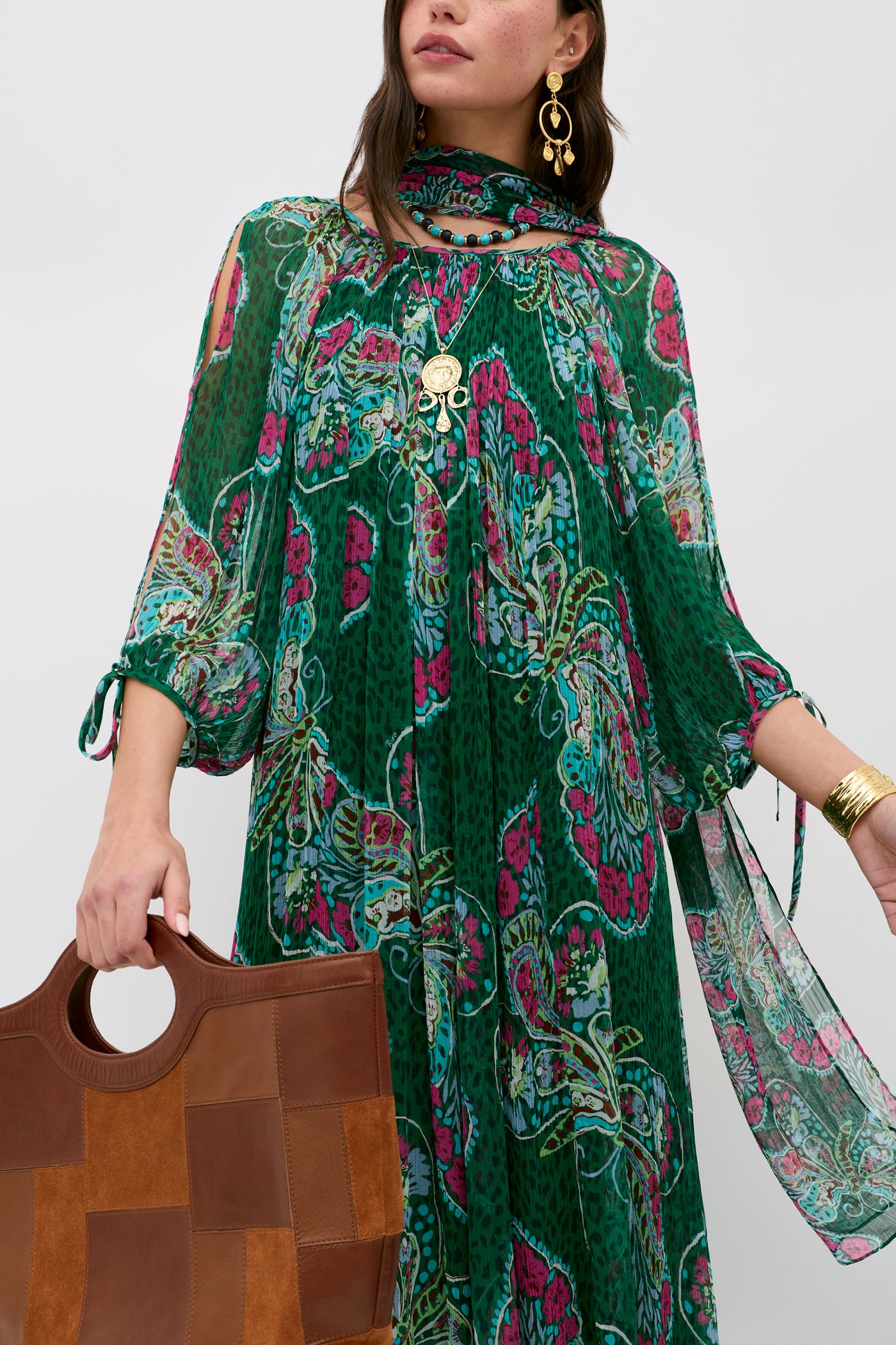 A woman wears the Hayden - Butterfly Bloom Green dress and carries a brown patchwork handbag, accessorized with gold jewelry—earrings, necklace, rings, and bracelet. Her face is partially out of frame.