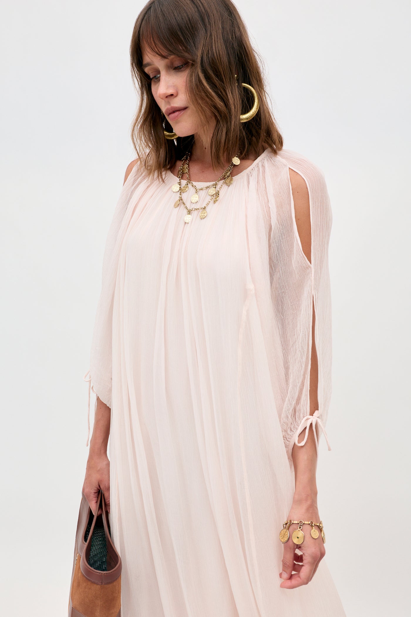 A woman wearing the Hayden - Shell light, flowy blush pink dress with shoulder cutouts, styled with gold hoop earrings, a layered necklace, and bracelet, holds a brown clutch against a plain background.