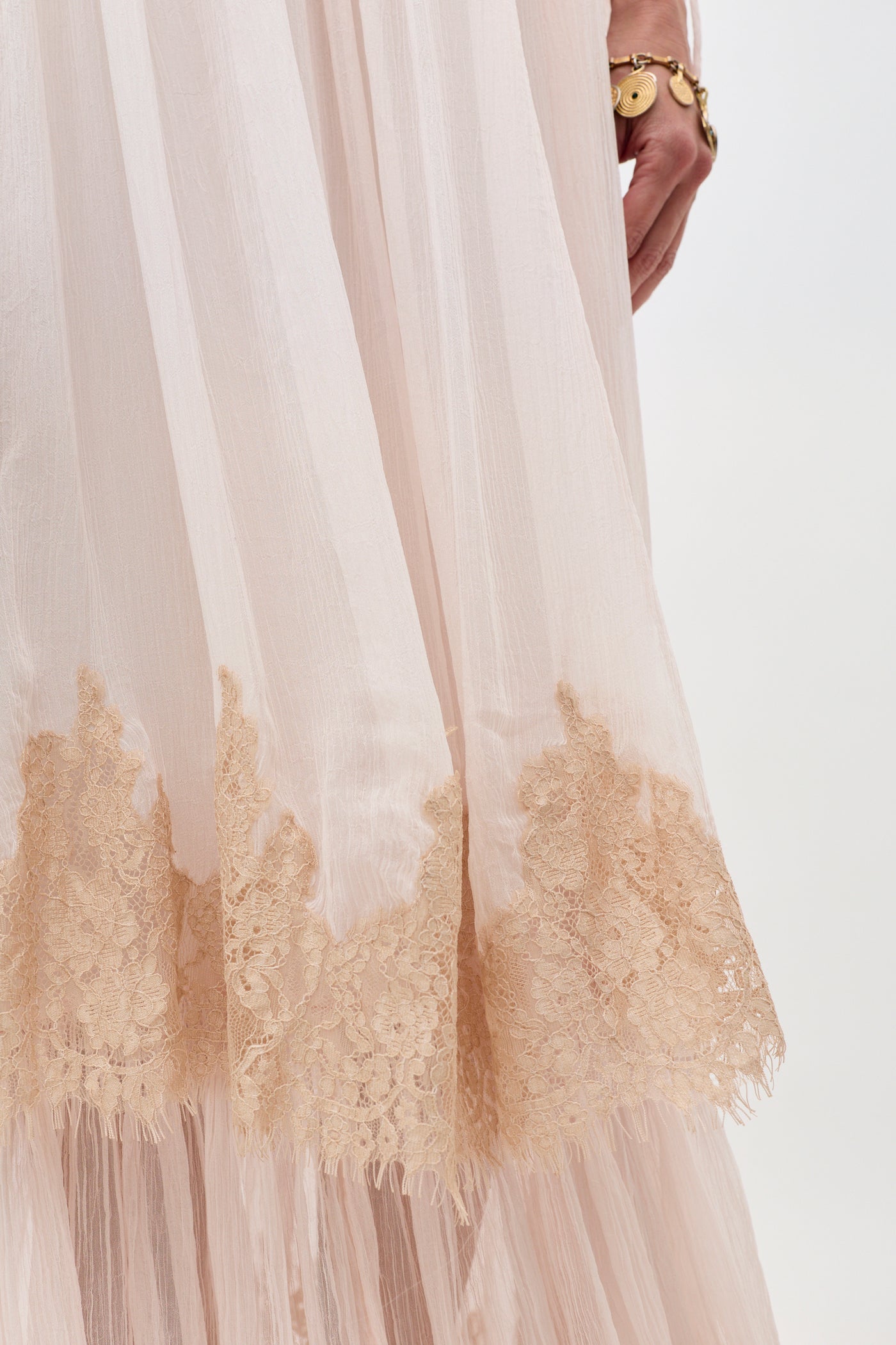 A close-up of the Hayden - Shell, a light pink sheer dress featuring beige lace trim near the hem. A person’s hand adorned with a gold bracelet appears on the right side of the image.