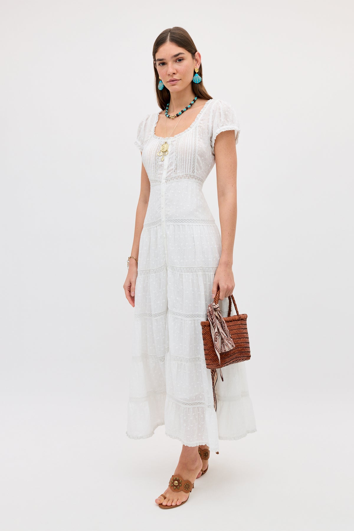 A woman with straight brown hair poses against a plain background, wearing the Jaclyn - Ivory cotton summer dress with lace details. She accessorizes with a brown woven handbag, tan sandals, turquoise earrings, and a necklace.
