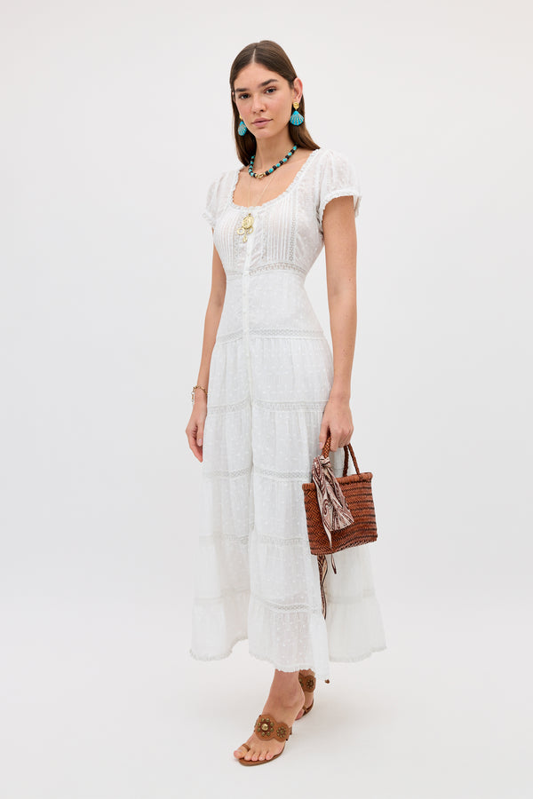 A woman with straight brown hair poses against a plain background, wearing the Jaclyn - Ivory cotton summer dress with lace details. She accessorizes with a brown woven handbag, tan sandals, turquoise earrings, and a necklace.