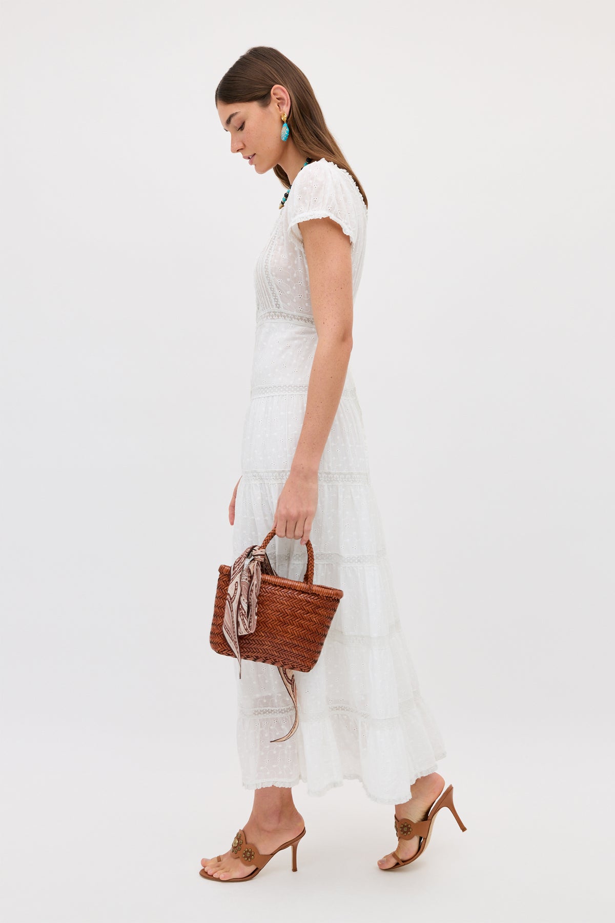 A woman wears the Jaclyn - Ivory cotton summer dress, holding a woven brown handbag with a scarf on the handle. She pairs it with tan heeled sandals and turquoise earrings, standing against a simple light background.