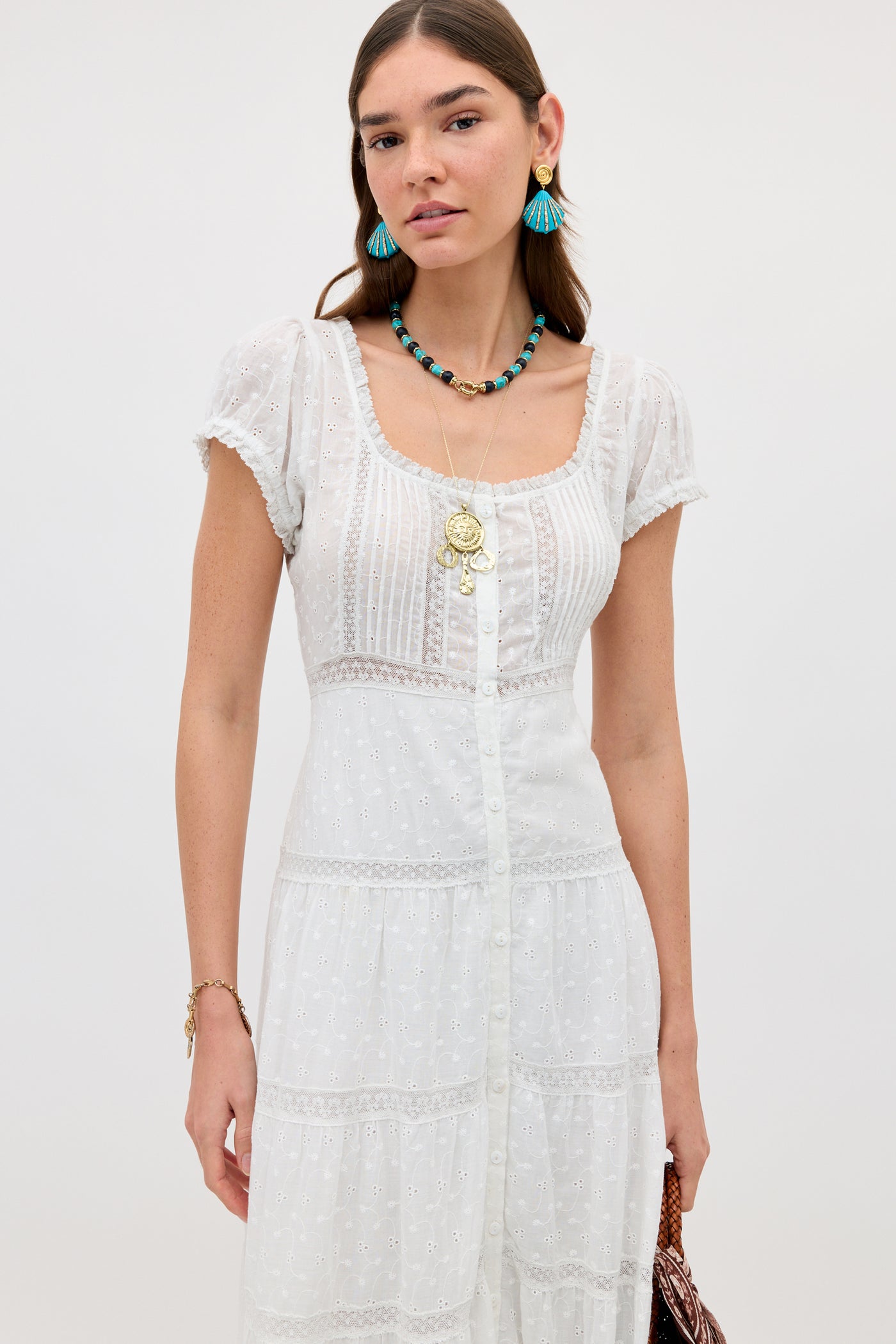 A woman stands against a plain background wearing the Jaclyn - Ivory dress, featuring white cotton broderie anglaise and short sleeves. She pairs it with colorful earrings, layered necklaces, and a bracelet.