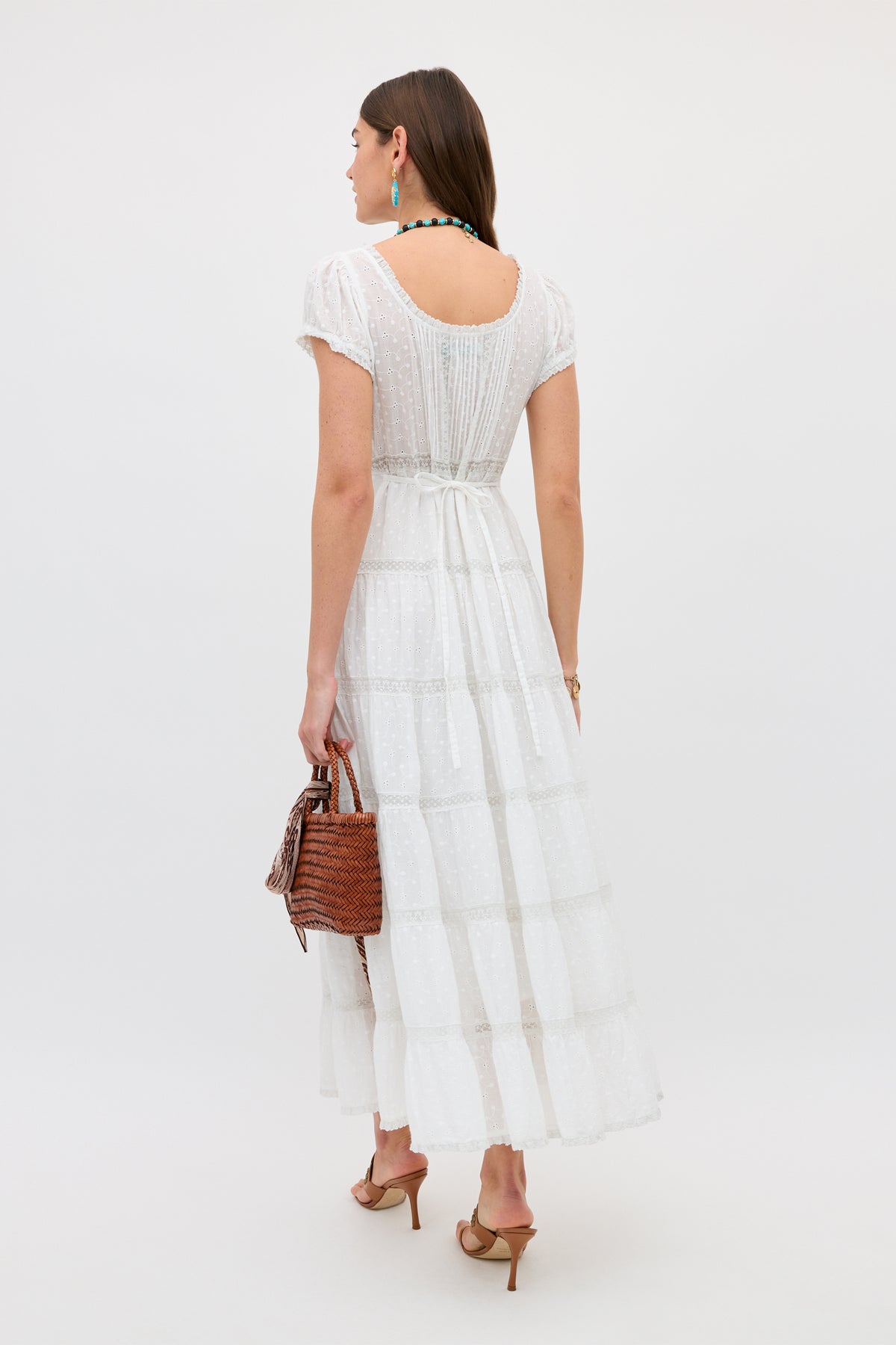 A woman in the Jaclyn - Ivory dress faces away, holding a brown woven handbag. She wears heeled sandals, a necklace, earrings, and has her brown hair down against a plain light background.