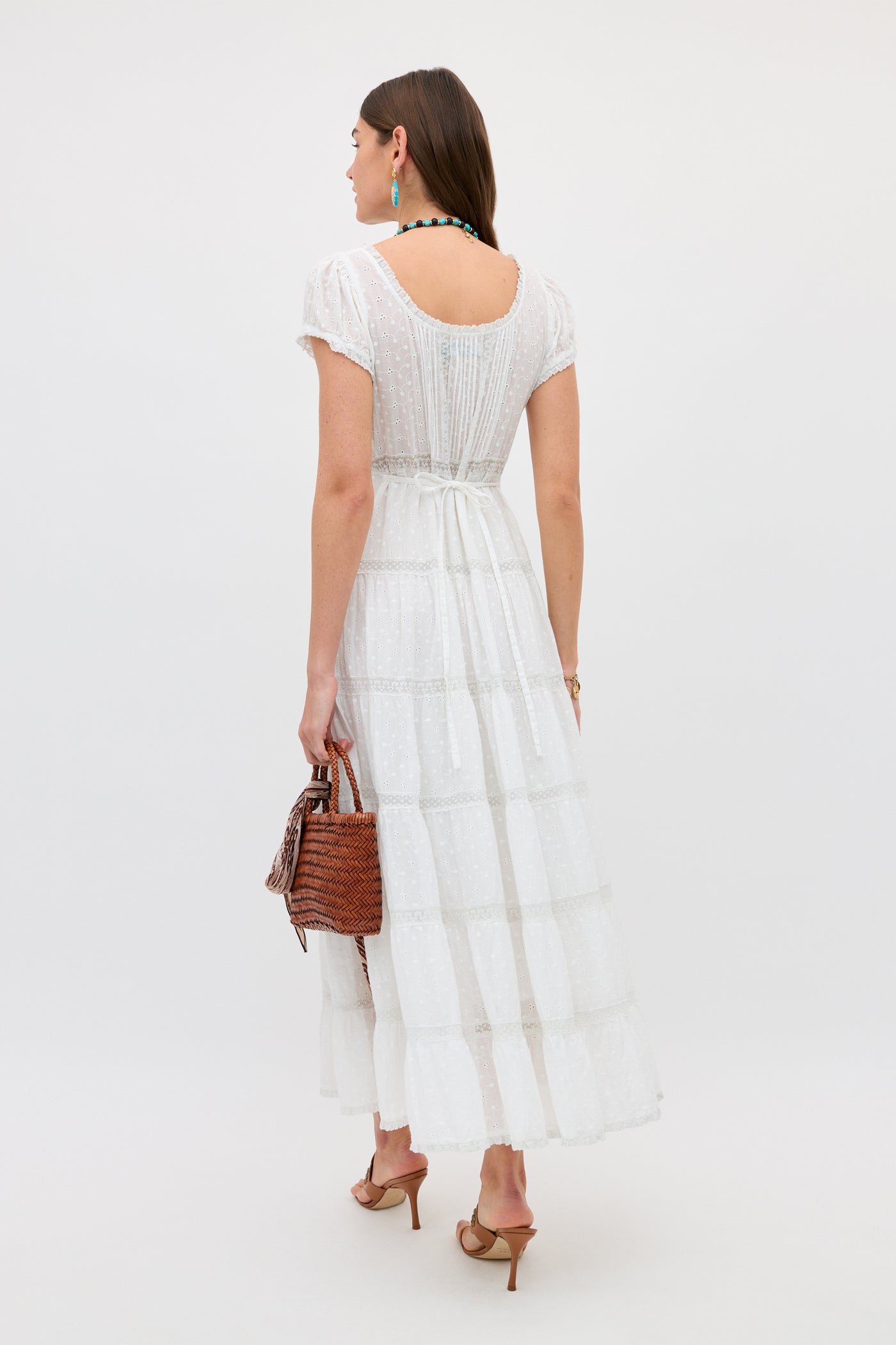 A woman in the Jaclyn - Ivory dress faces away, holding a brown woven handbag. She wears heeled sandals, a necklace, earrings, and has her brown hair down against a plain light background.