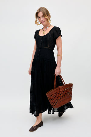 A woman wears the Jaclyn - Black short-sleeve, button-front maxi dress, holding a brown woven tote bag and wearing matching flats, necklace, and bracelet against a plain light background.