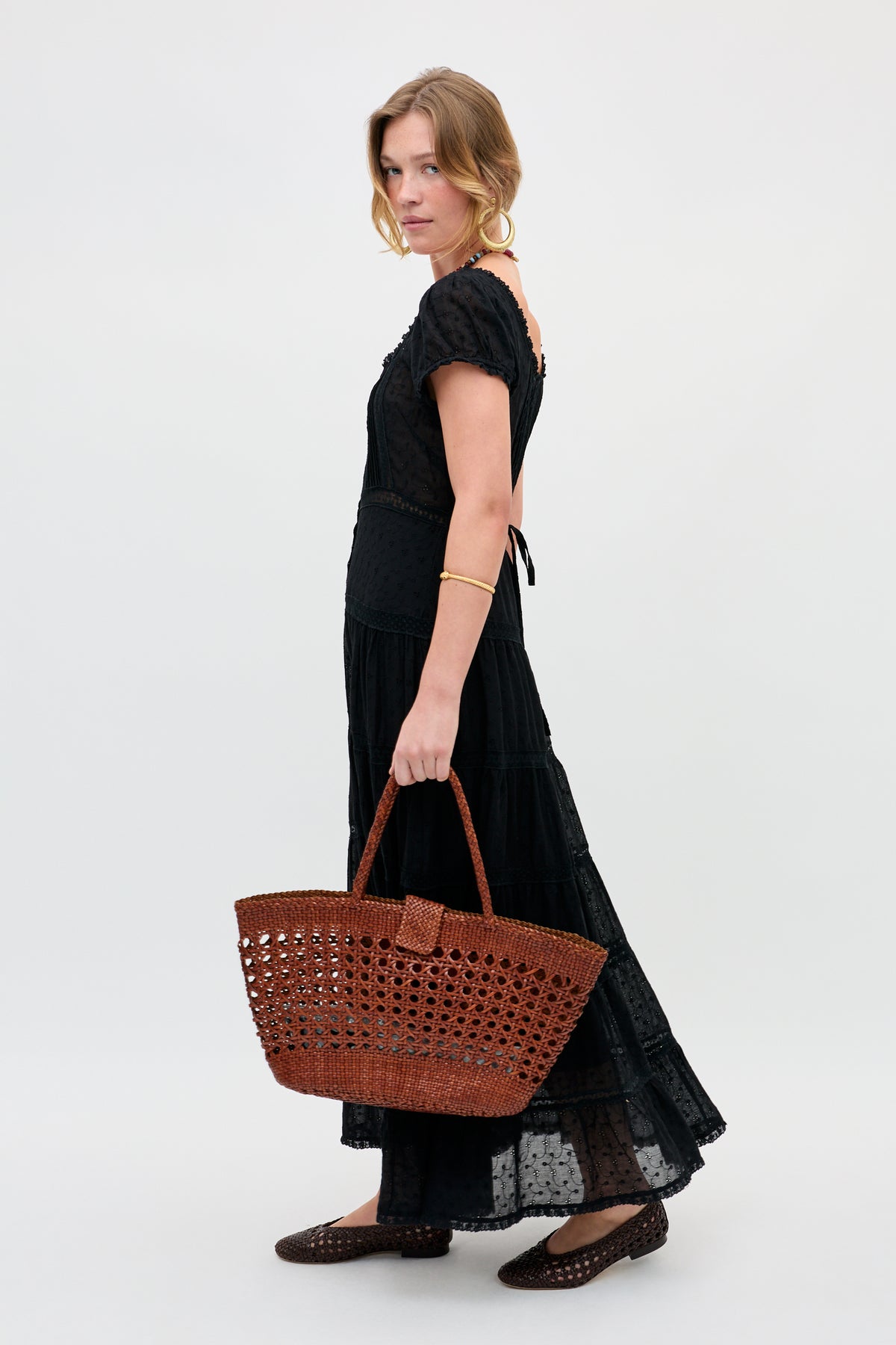 A woman with loosely styled blonde hair looks over her shoulder at the camera, wearing the Jaclyn - Black dress and black woven shoes, and holding a large brown woven handbag.