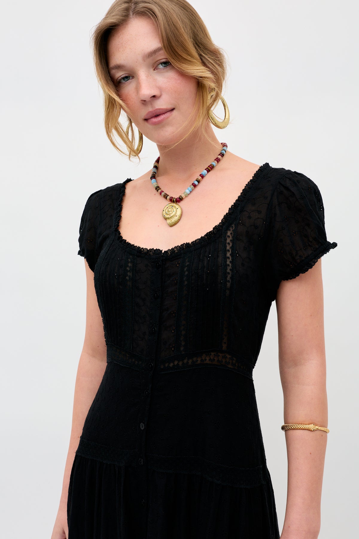 A light-skinned blonde woman in loose waves wears the Parma - Multi: a black, short-sleeved textured dress, colorful beaded necklace with gold pendant, gold hoop earrings, and a gold bracelet against a plain background.