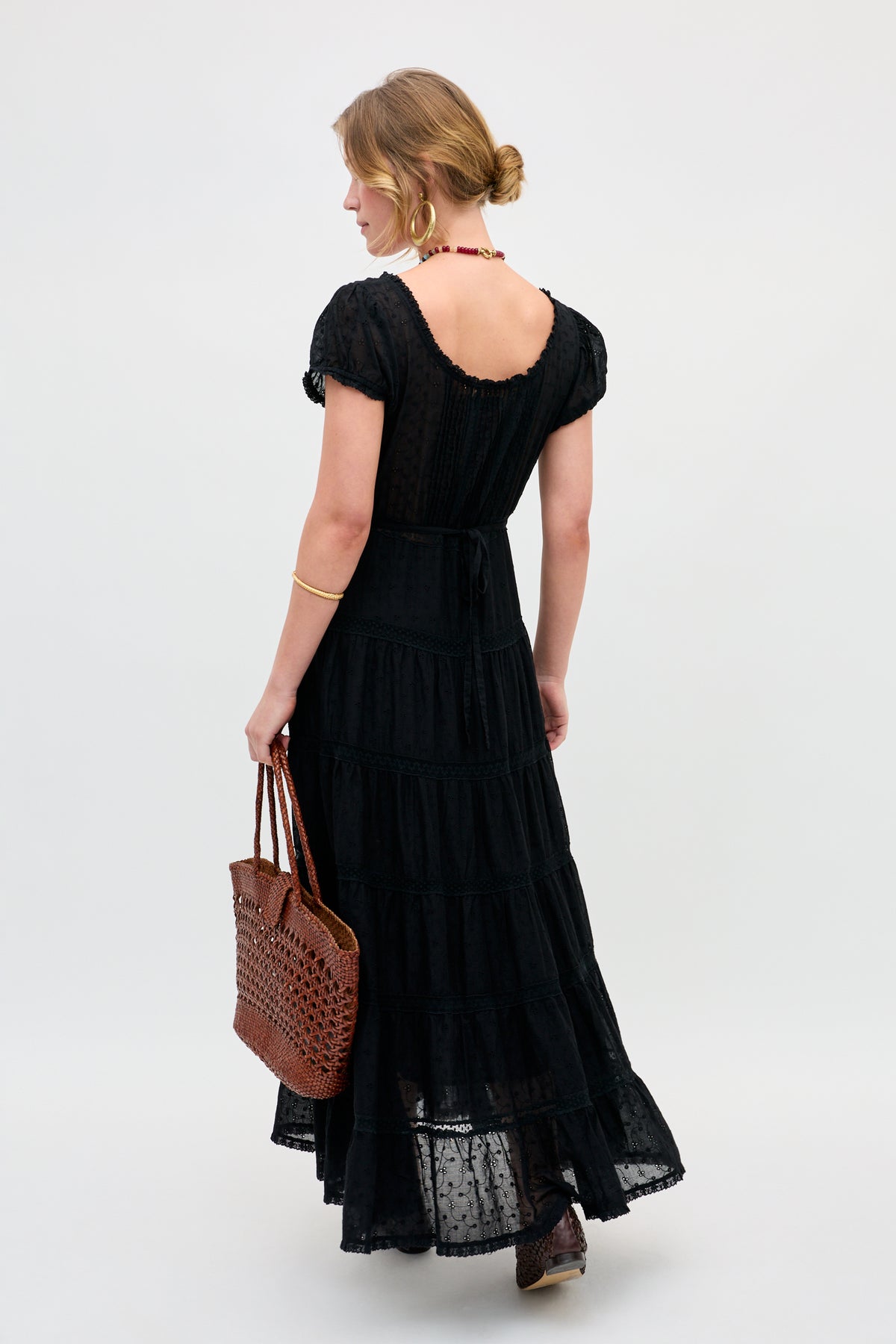 A woman with blonde hair in a low bun, facing away against a light background, wears the Jaclyn - Black long dress with short sleeves and holds a brown woven tote bag.