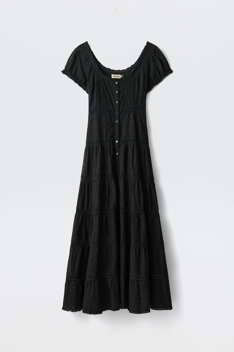 The Jaclyn - Black is an ankle-length, short-sleeve dress with a scoop neckline, button-down front, fitted bodice, flared tiered skirt, and subtle texture details. Shown against a plain white background.