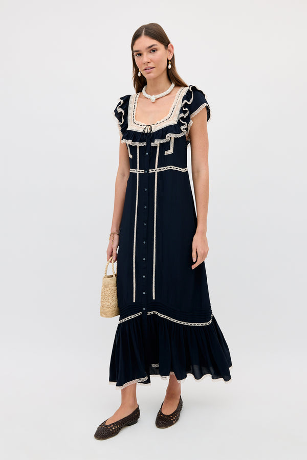 A woman poses against a plain background in the Kendara - Deep Navy dress, featuring white trim and ruffle accents. She carries a small woven bag, wears dark flats, has her hair down, and accessorizes with jewelry.