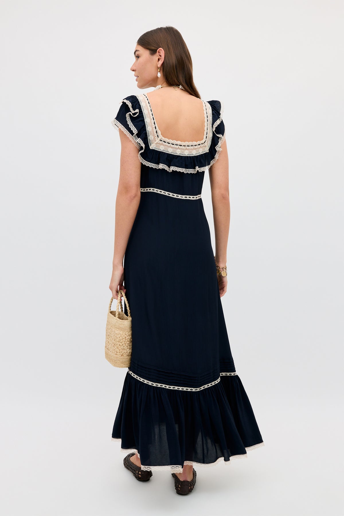 A woman with long brown hair wears the Kendara - Deep Navy, a prairie-inspired ankle-length cotton dress with ruffled sleeves, lace trim, and square low back, holding a woven straw handbag and facing away.