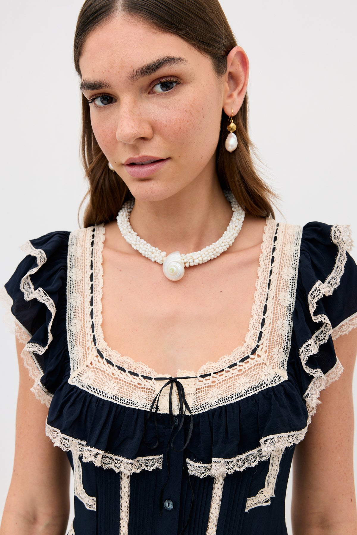 A woman with straight brown hair wears the Kendara - Deep Navy cotton midi dress with lace trim, a multi-strand pearl necklace featuring a shell pendant, and pearl drop earrings. She looks at the camera against a plain background.