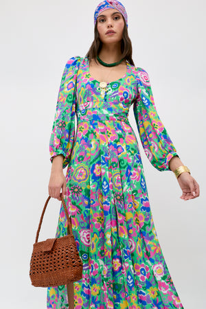 Wearing the Alfie - Ikat Bloom Green dress, a woman pairs its vibrant floral design and puffed sleeves with a purple headscarf, gold jewelry, a green necklace, and a woven brown handbag against a light background.