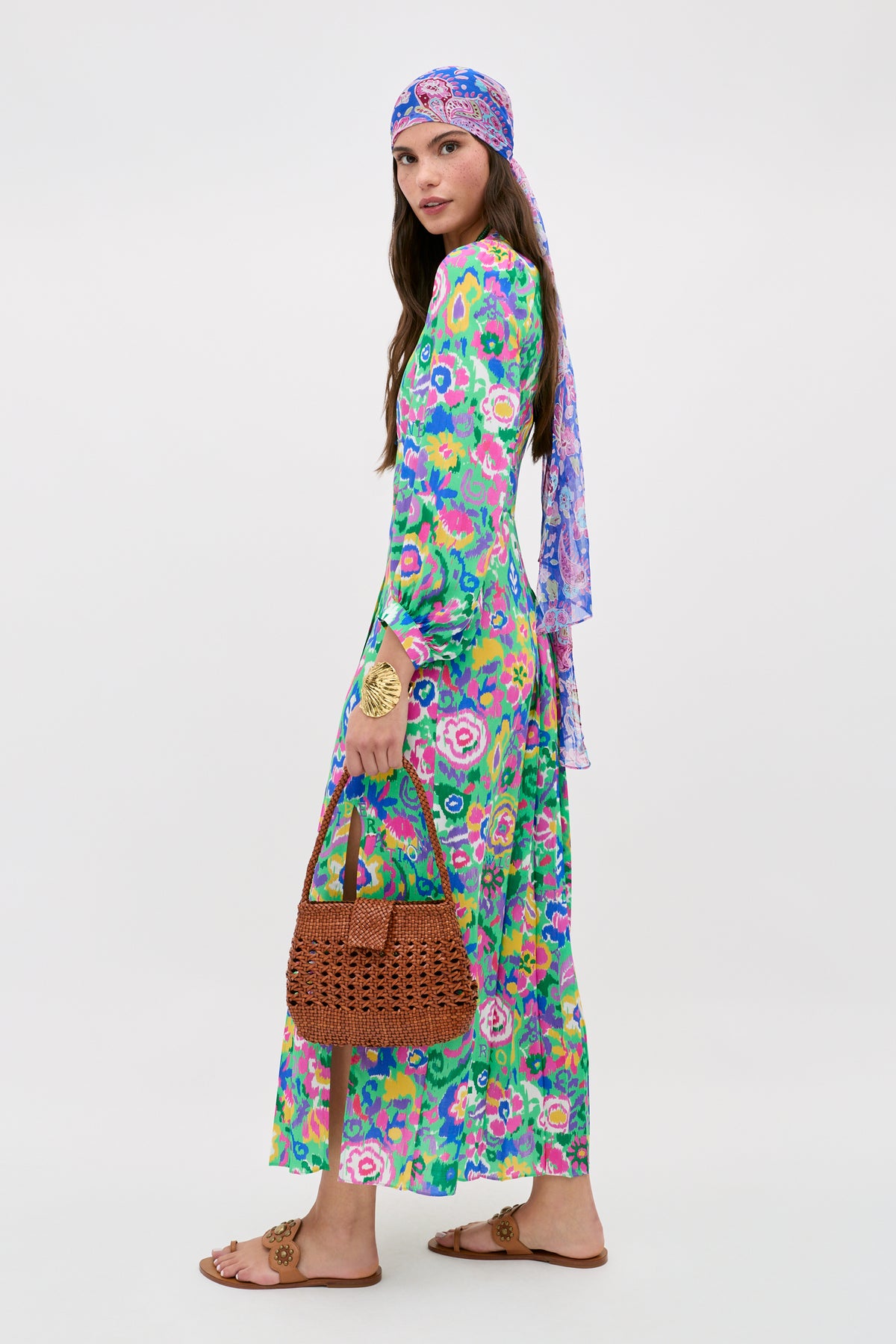 A woman in the "Alfie - Ikat Bloom Green" floral maxi dress with a matching headscarf and sandals stands sideways, holding a brown woven handbag and wearing a large gold bracelet against a plain white background.