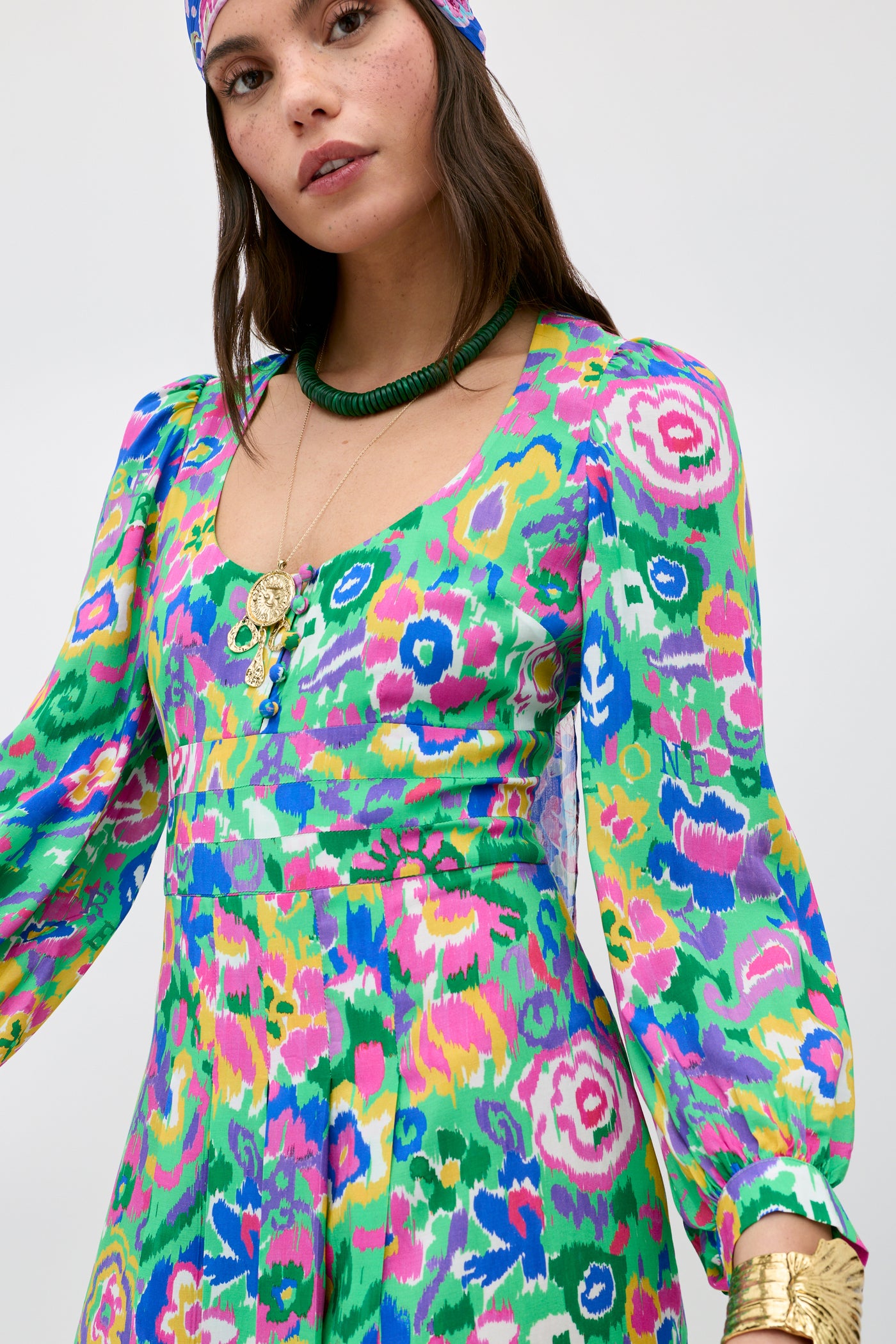 A woman wearing the Alfie - Ikat Bloom Green patterned dress with long sleeves, layered gold necklaces, a green beaded necklace, matching headscarf, and gold bracelets stands against a plain light background.
