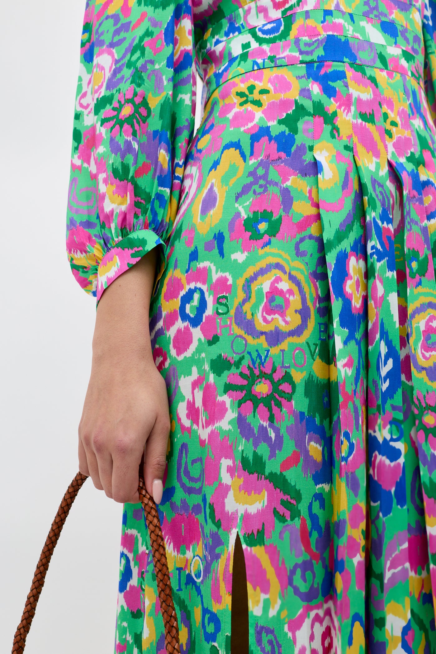 Wearing the Alfie - Ikat Bloom Green dress featuring a colorful floral print, pleats, and a front slit, she carries a brown braided handle bag.