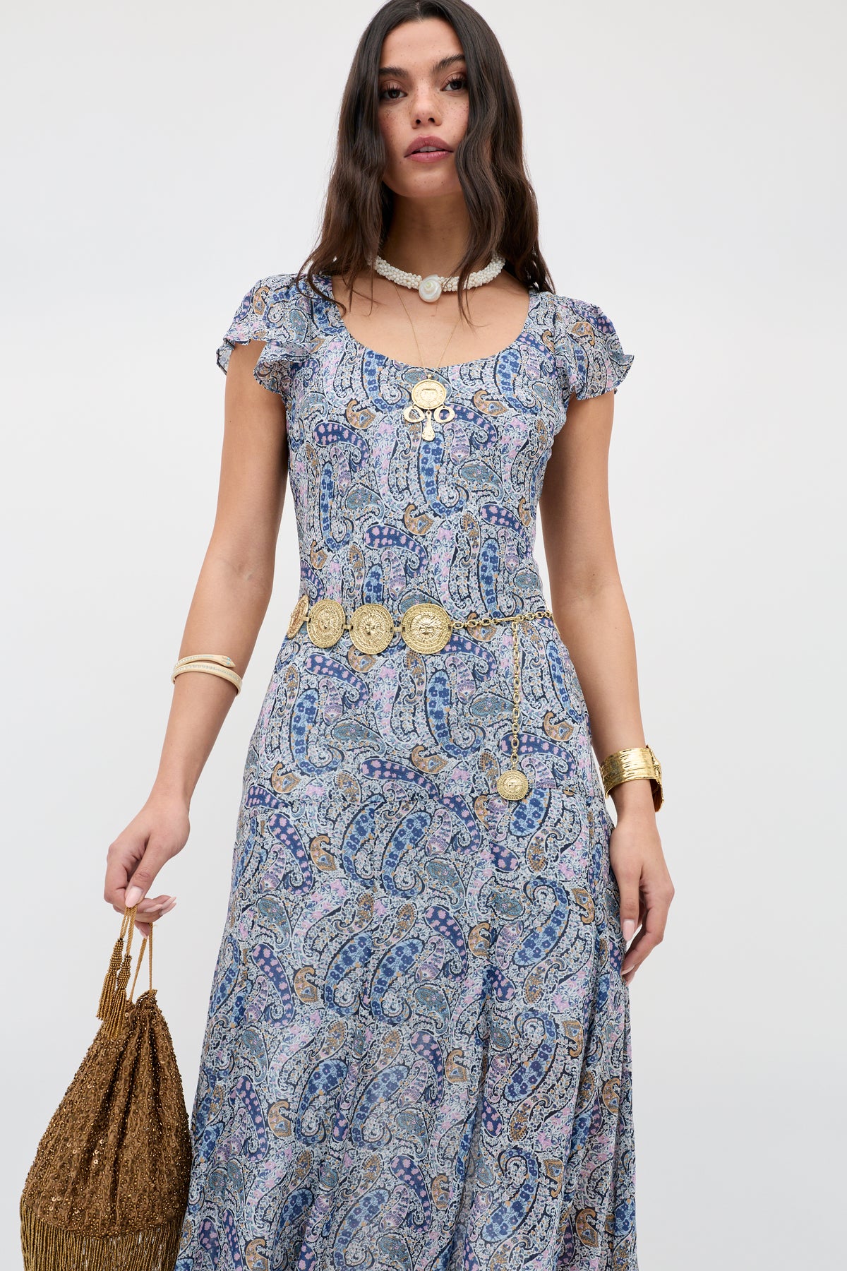 Woman wearing Leyla - Gold, a blue and white paisley dress with layered gold jewelry and a decorative gold belt, holding a brown woven bag against a plain light background.