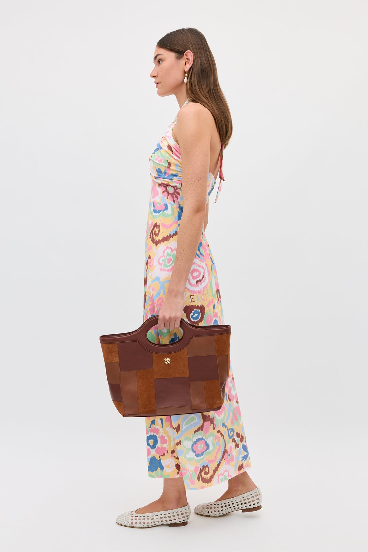 A woman in the Ilyana - Ikat Bloom Yellow dress stands sideways, holding a large brown tote with green handles. She pairs it with white woven flats and drop earrings against a plain light background.