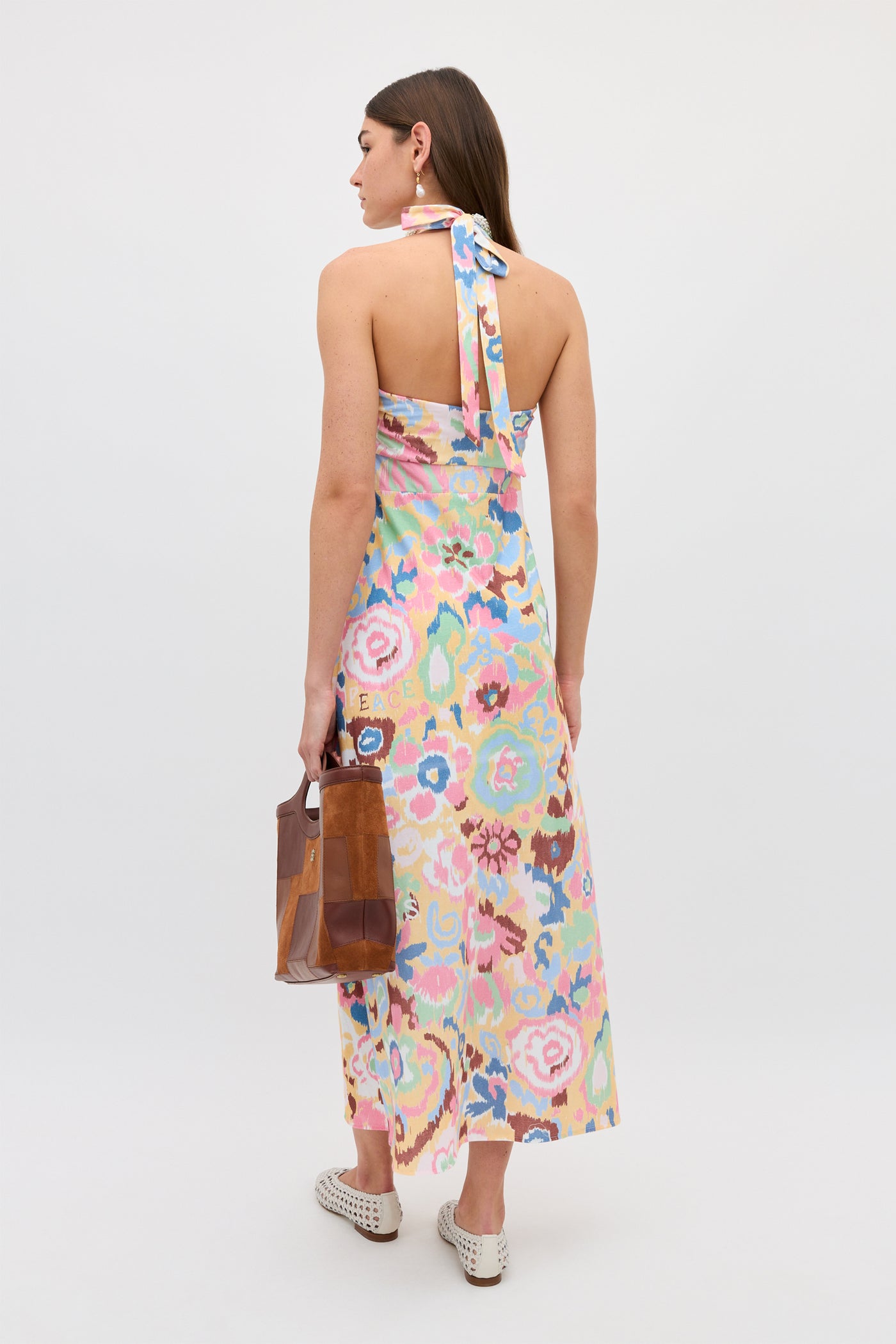 A woman faces away, wearing the Ilyana - Ikat Bloom Yellow floral halter dress. She holds a brown patchwork handbag and wears white woven slip-on shoes, all set against a plain white background.