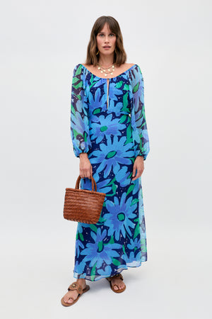 A woman stands against a plain white background wearing the Nikolette - Danish Floral Blue long, blue floral dress with long sleeves, paired with brown sandals, a chunky gold necklace, and holding a woven brown handbag.