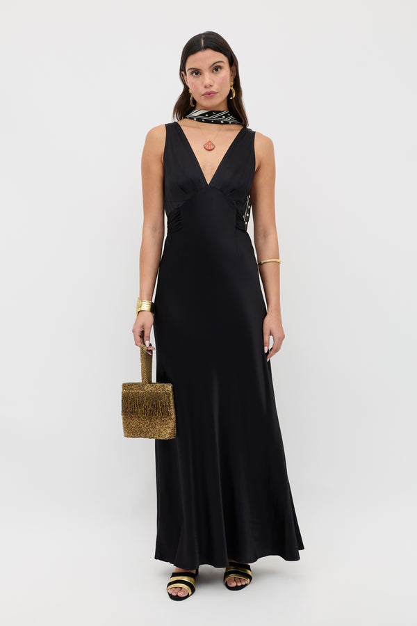 A woman stands against a plain white background wearing the Evalise - Black sleeveless deep V-neck satin dress, gold and black sandals, gold jewelry, a neck scarf, and holding a textured brown handbag.