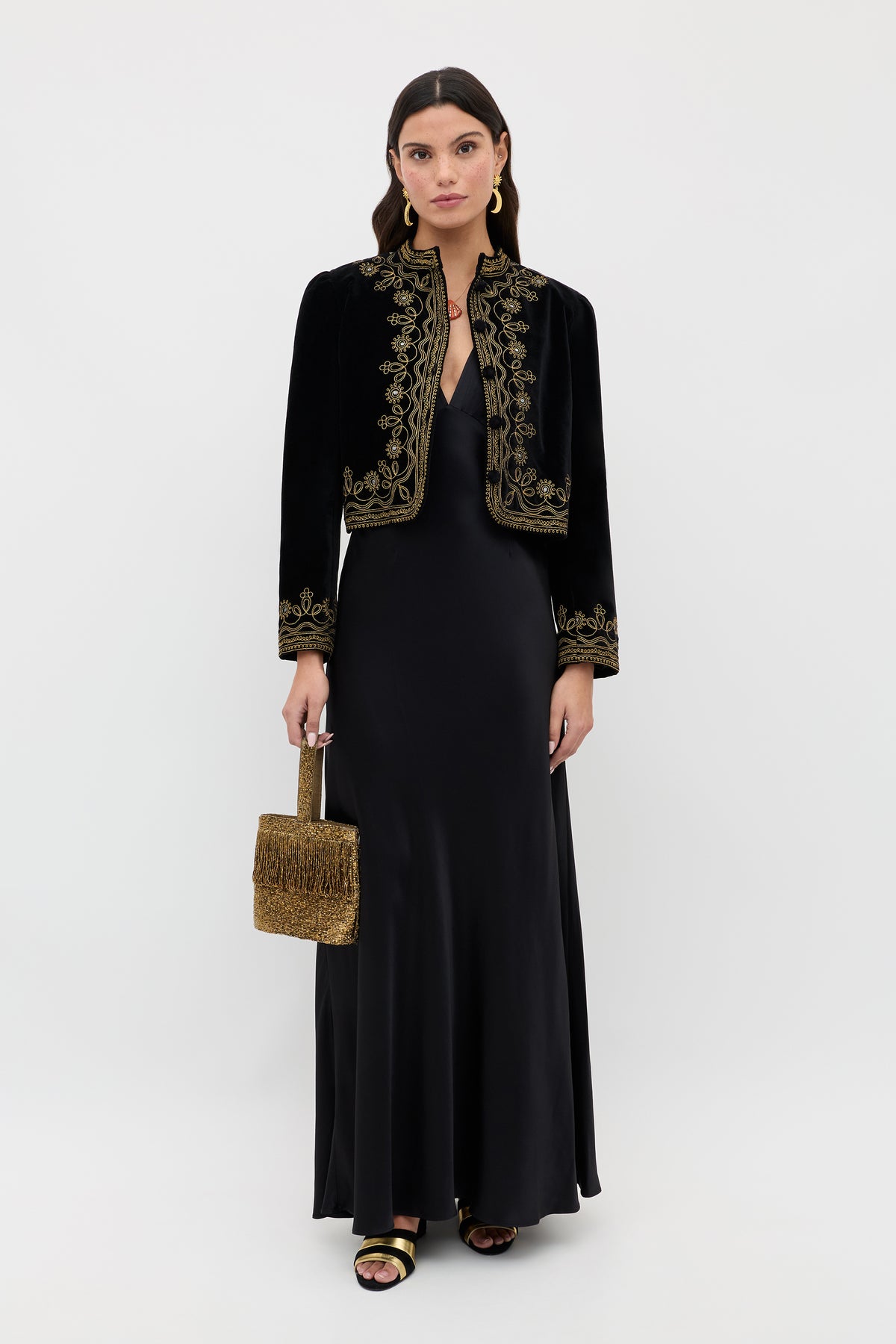 A woman stands against a white background wearing the Evalise - Black, a long black satin dress paired with a black velvet jacket with gold embroidery, black and gold sandals, and carrying a small gold textured handbag.
