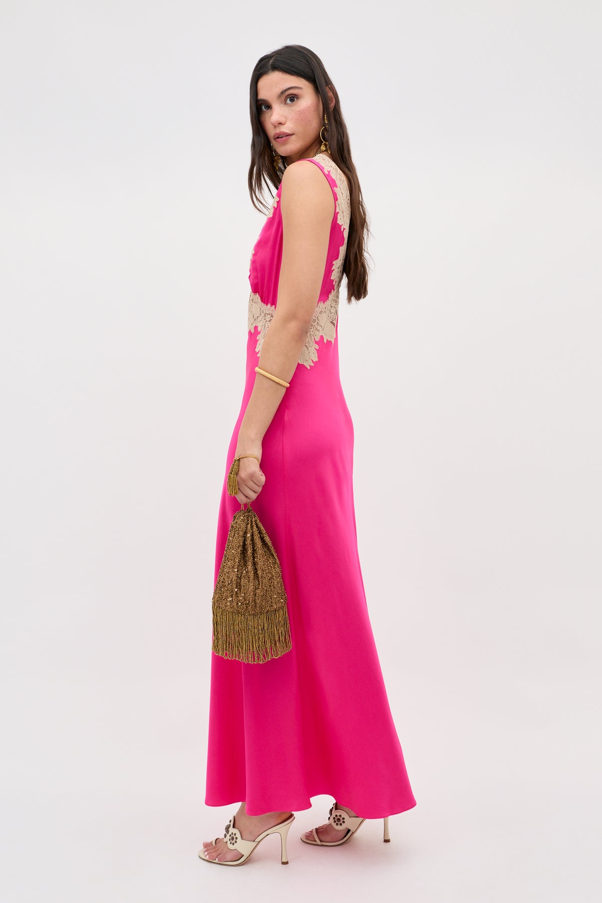 A woman wears the Lysandra - Cerise, a sleeveless, floor-length hot pink dress with lace details. She accessorizes with gold earrings, heeled sandals, and a gold fringed handbag, posing against a plain white background.