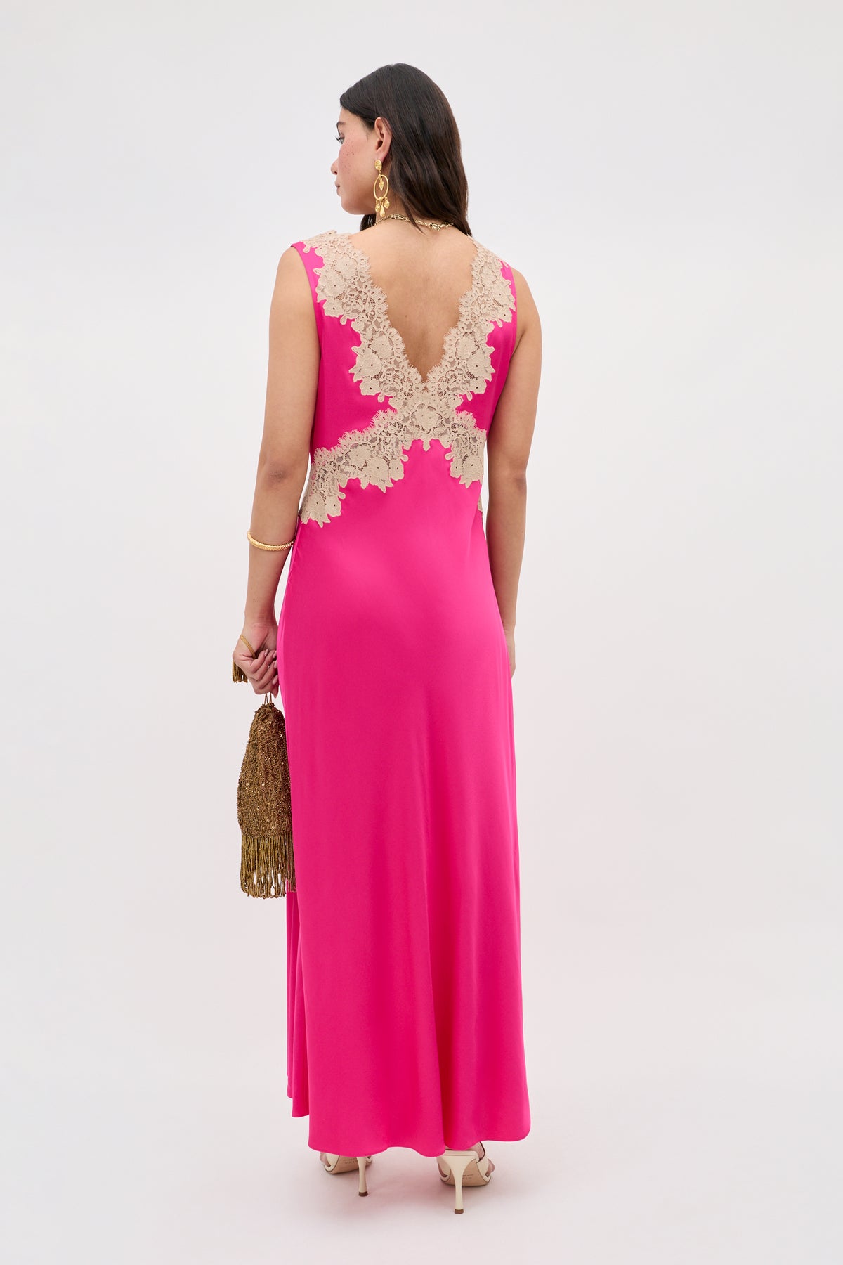 A woman faces away in the Lysandra - Cerise, a sleeveless floor-length hot pink dress with beige lace on the upper back. She holds a gold tasseled purse and wears gold earrings and heels against a plain background.