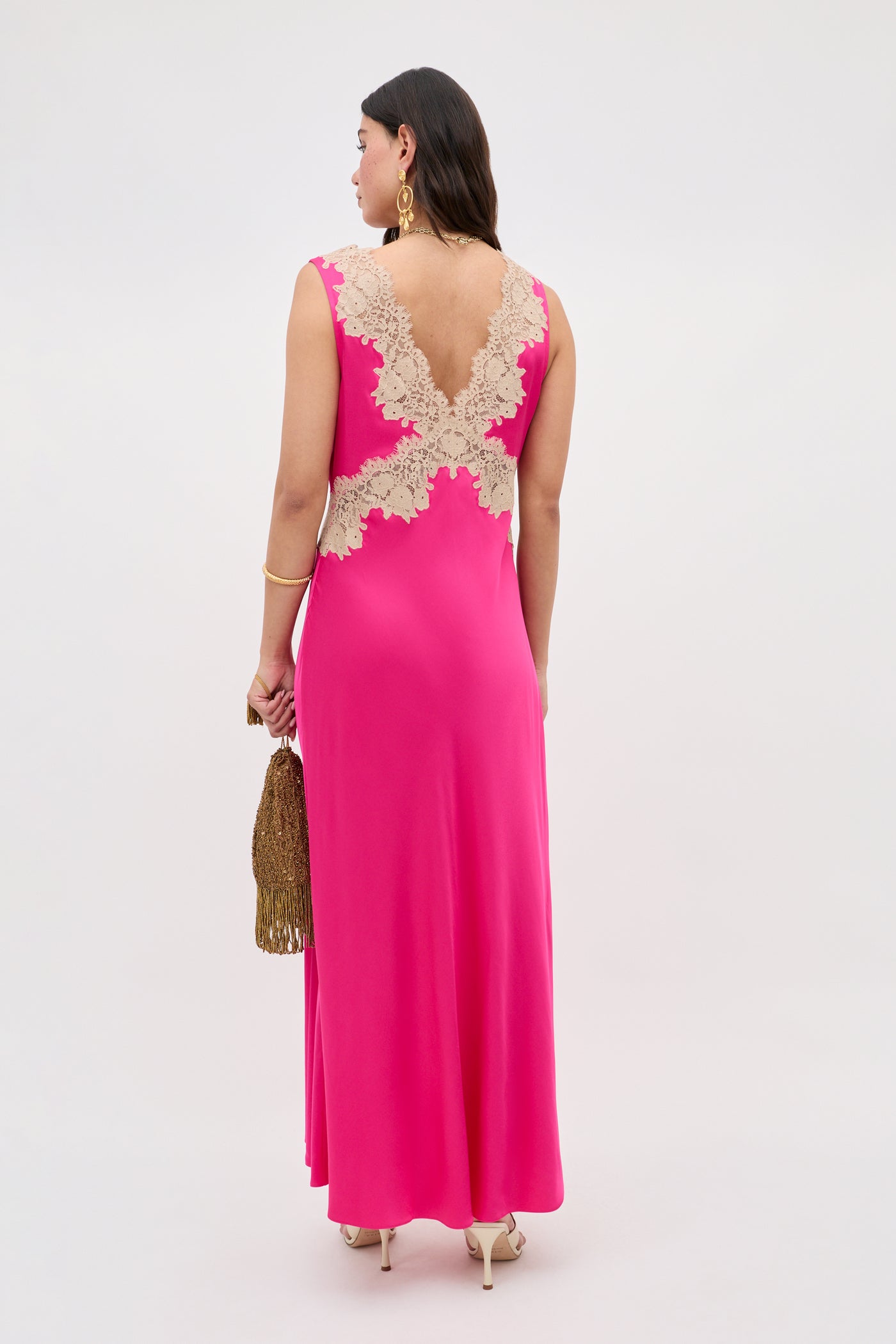 A woman faces away in the Lysandra - Cerise, a sleeveless floor-length hot pink dress with beige lace on the upper back. She holds a gold tasseled purse and wears gold earrings and heels against a plain background.