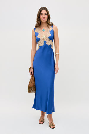 A woman stands against a plain light background wearing the Lysandra - Iris Blue sleeveless satin dress with beige lace bodice, holding a brown fringe handbag, paired with strappy heels and gold jewelry.