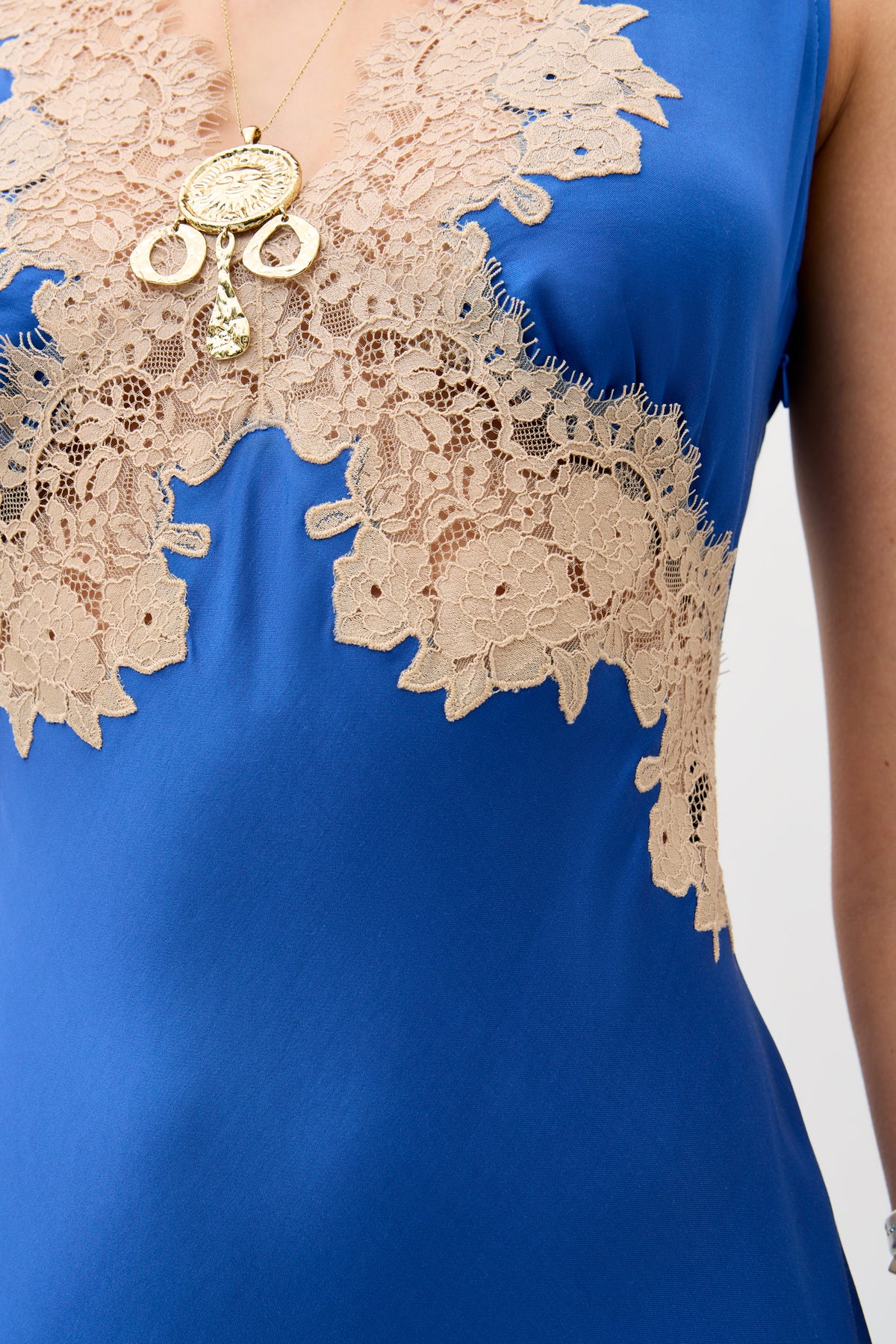 Close-up of a person wearing the Lysandra - Iris Blue dress with beige lace around the neckline and chest, paired with a gold cameo pendant necklace featuring ornamental designs.