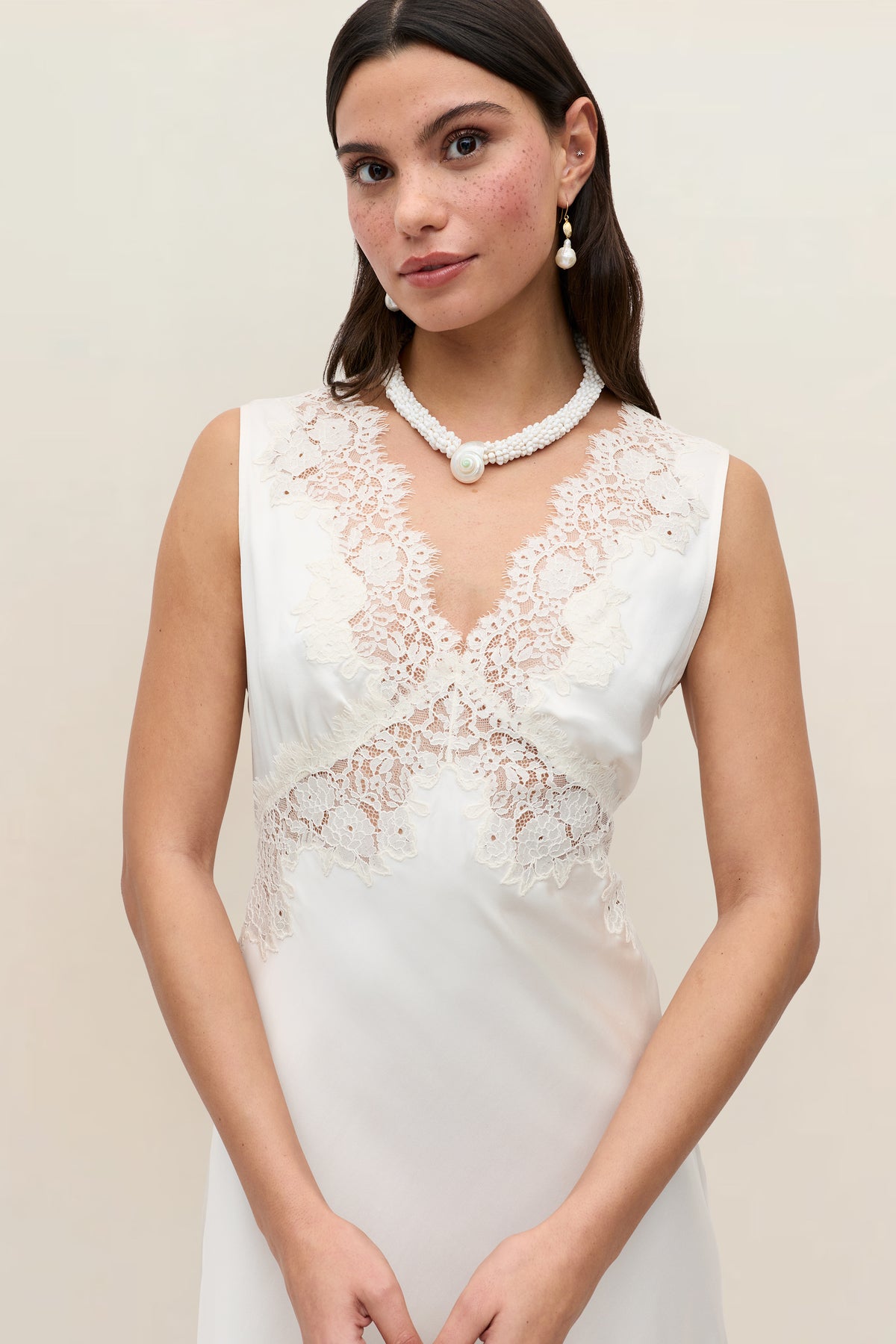 A woman in the Azzurra - Pearl sleeveless lace wedding dress poses against a plain background, accessorized with a white beaded necklace featuring a large pearl and matching pearl earrings. She has long dark hair.