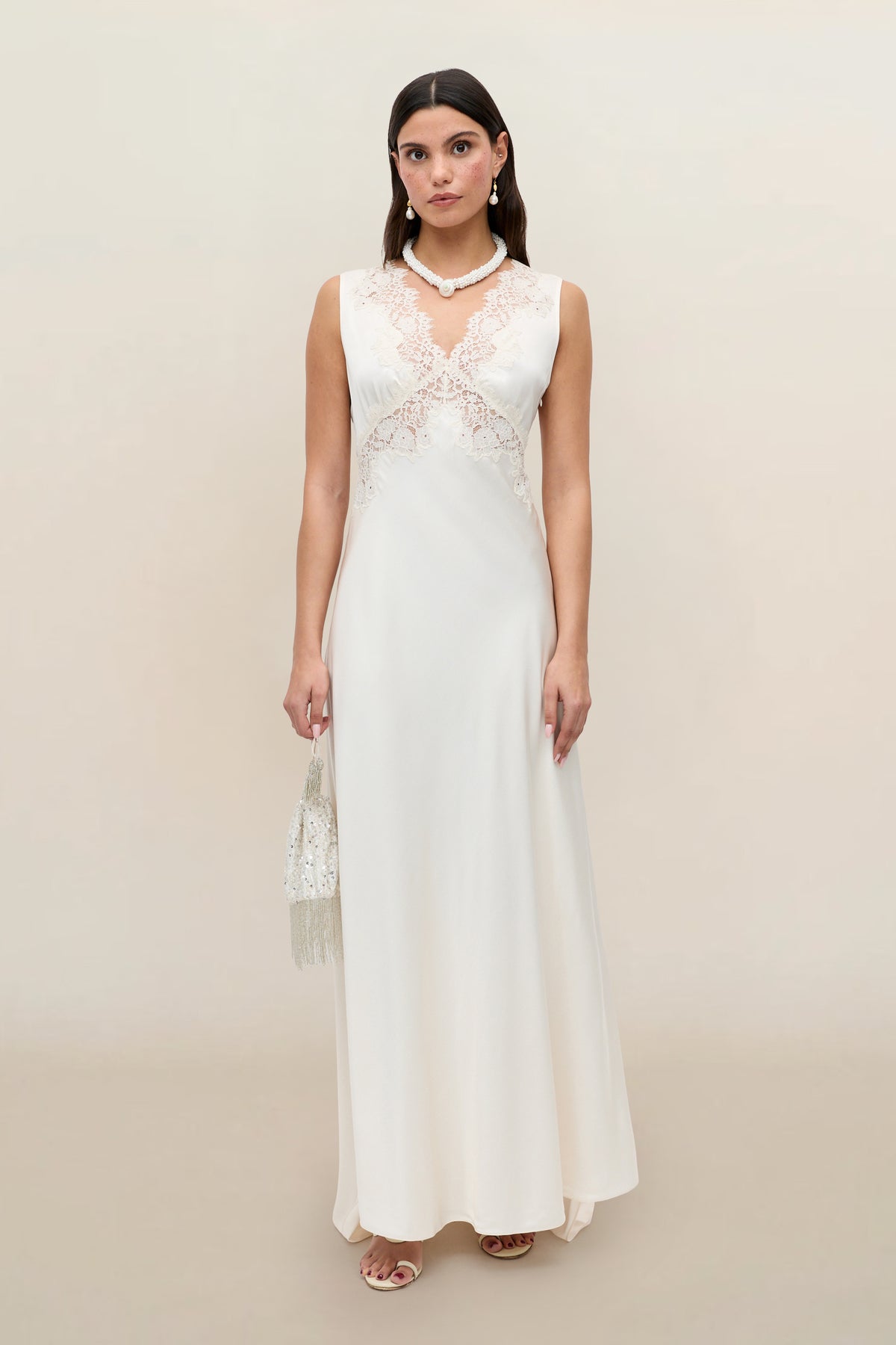 A woman in the Azzurra - Pearl sleeveless ivory lace wedding dress with a detailed bodice, pearl necklace and earrings, and a beaded handbag stands against a plain beige background.