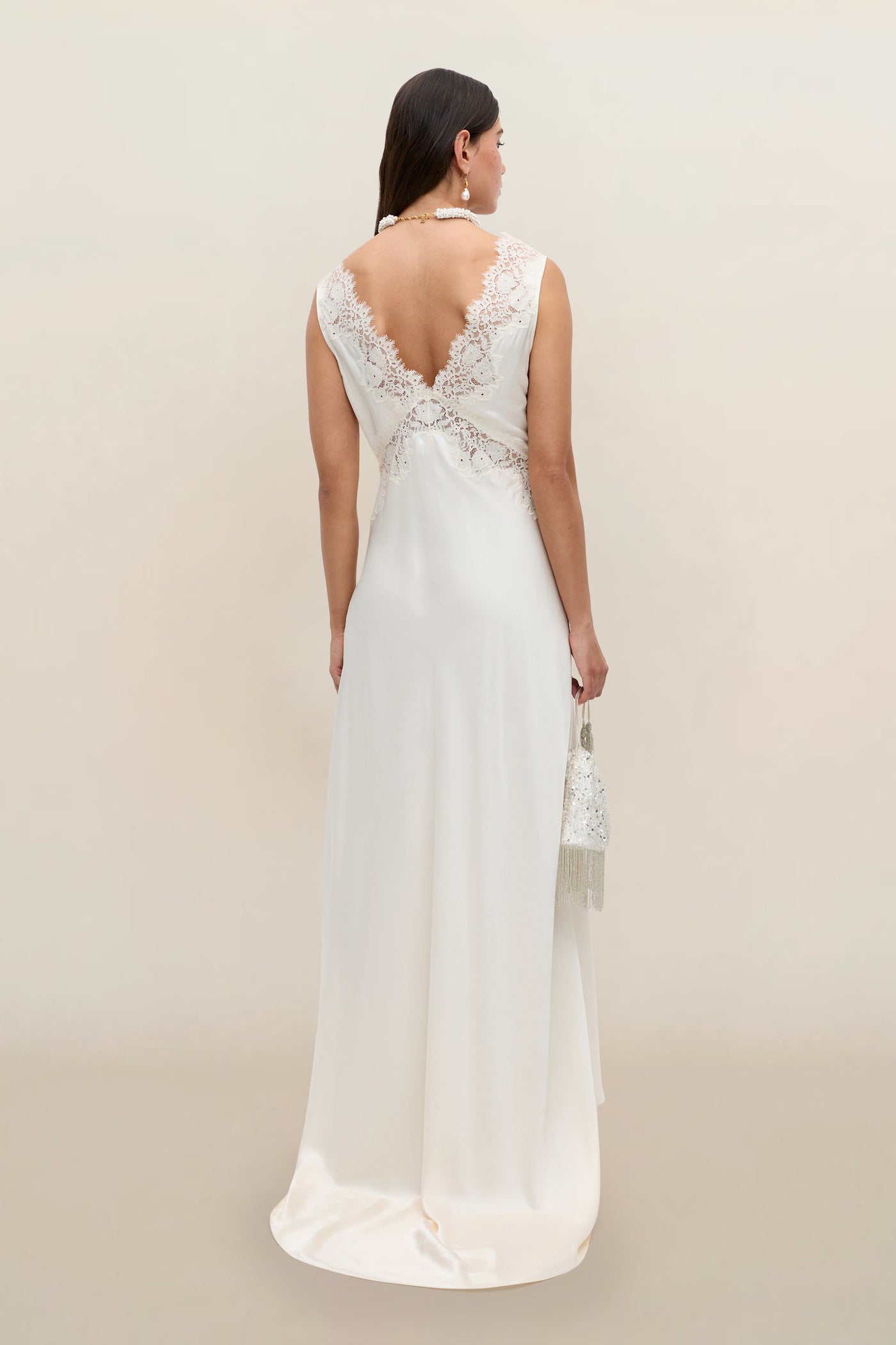 A woman in the Azzurra - Pearl sleeveless lace wedding dress with a deep V-back and flowing skirt stands facing away, holding a small beaded handbag. Delicate lace details complete this timeless, romantic bridal look.