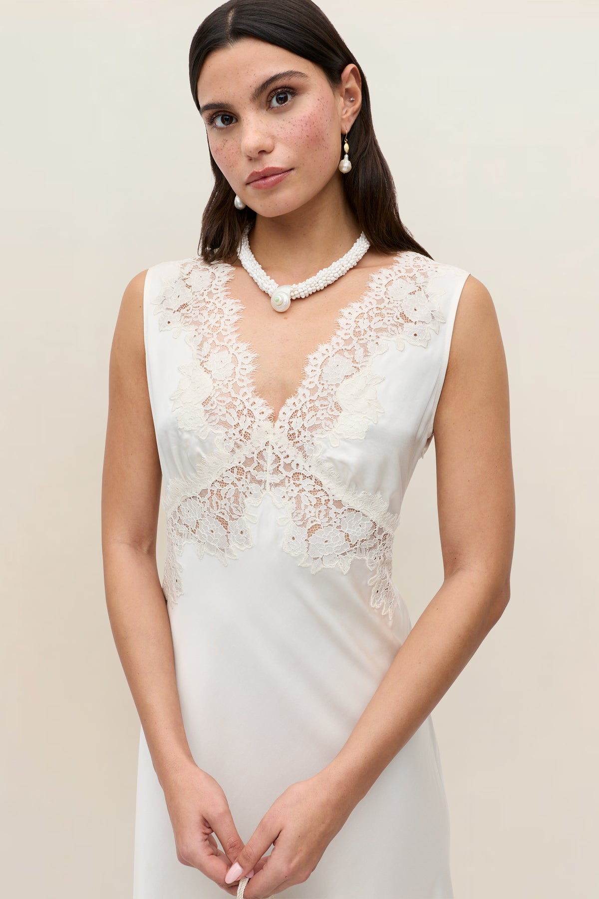 A woman wears the Azzurra – Pearl sleeveless white lace wedding dress with lace neckline, styled with pearl earrings and a matching necklace, standing against a plain light background.