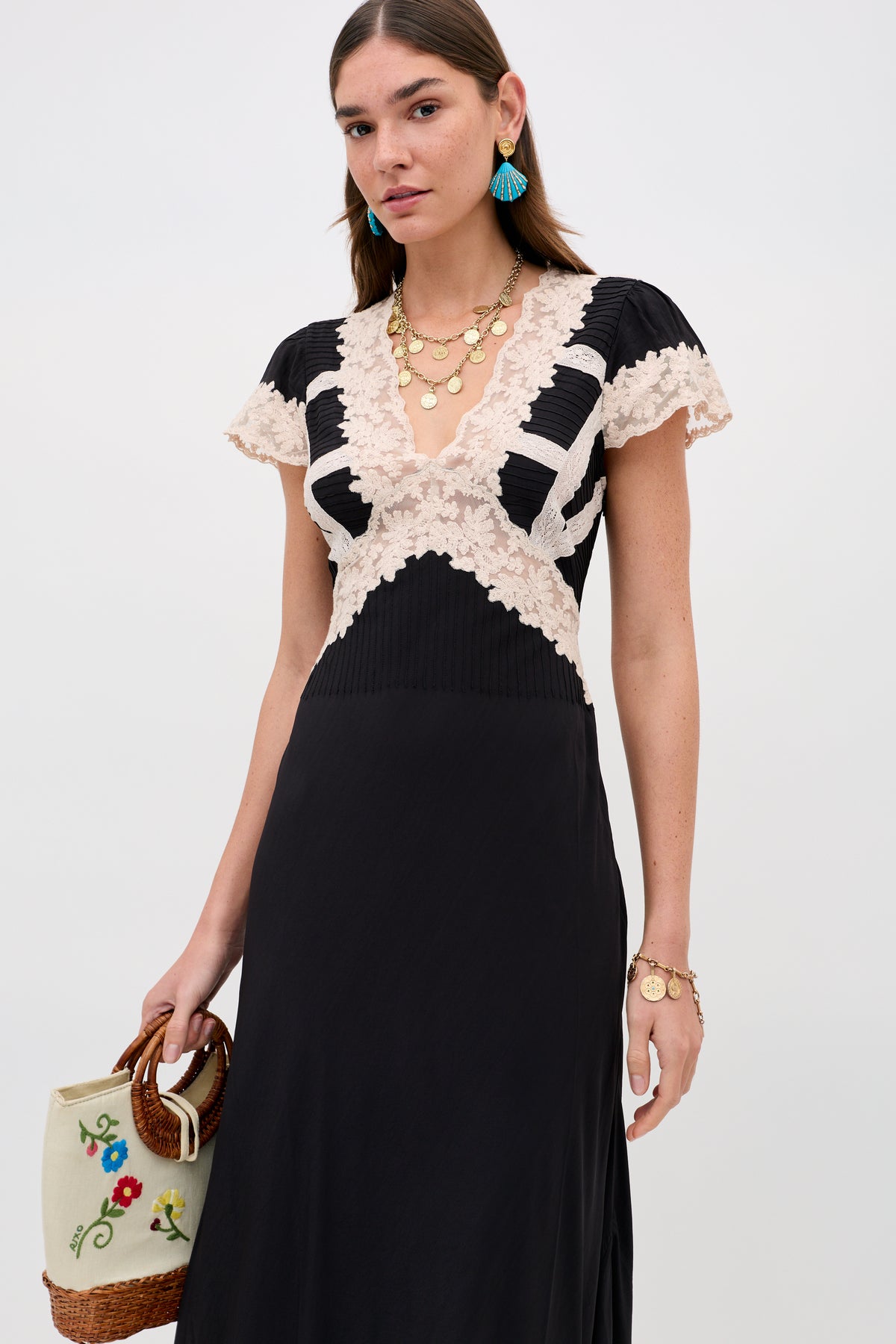A woman wearing the Martina - Black dress, layered gold necklaces, blue tassel earrings, and bracelets holds a cream handbag with colorful floral embroidery and a wicker handle against a plain white background.