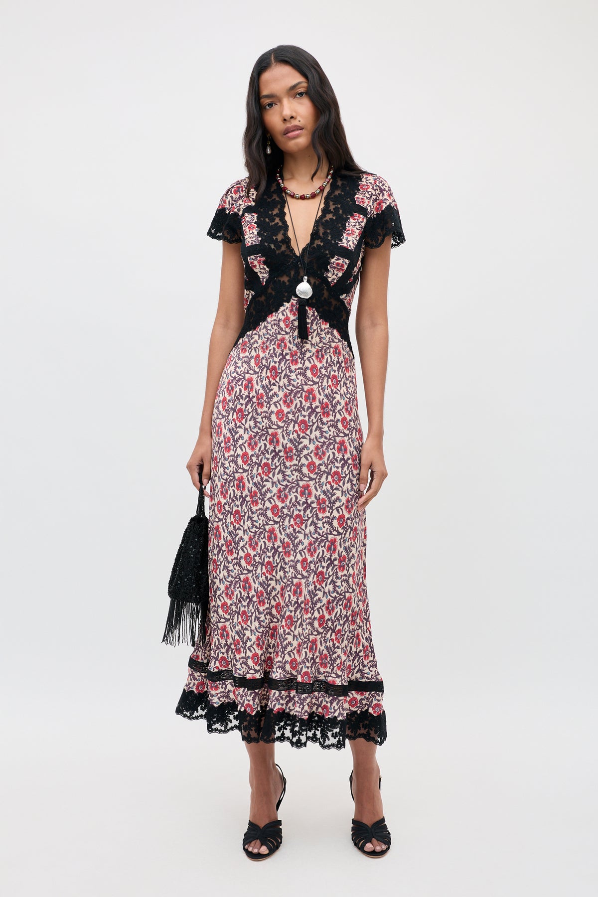 A woman wearing the Martina - Sarasa Rose Red floral midi dress with black lace trim, styled with black heels and a fringed black bag, stands against a plain light background.