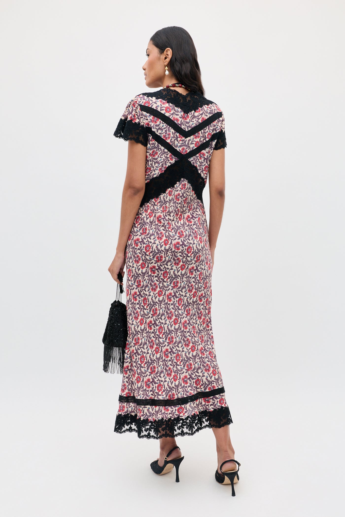A woman wearing the Martina - Sarasa Rose Red floral print dress with black lace trim, holding a black purse and wearing black high heels, stands facing away from the camera against a plain background.