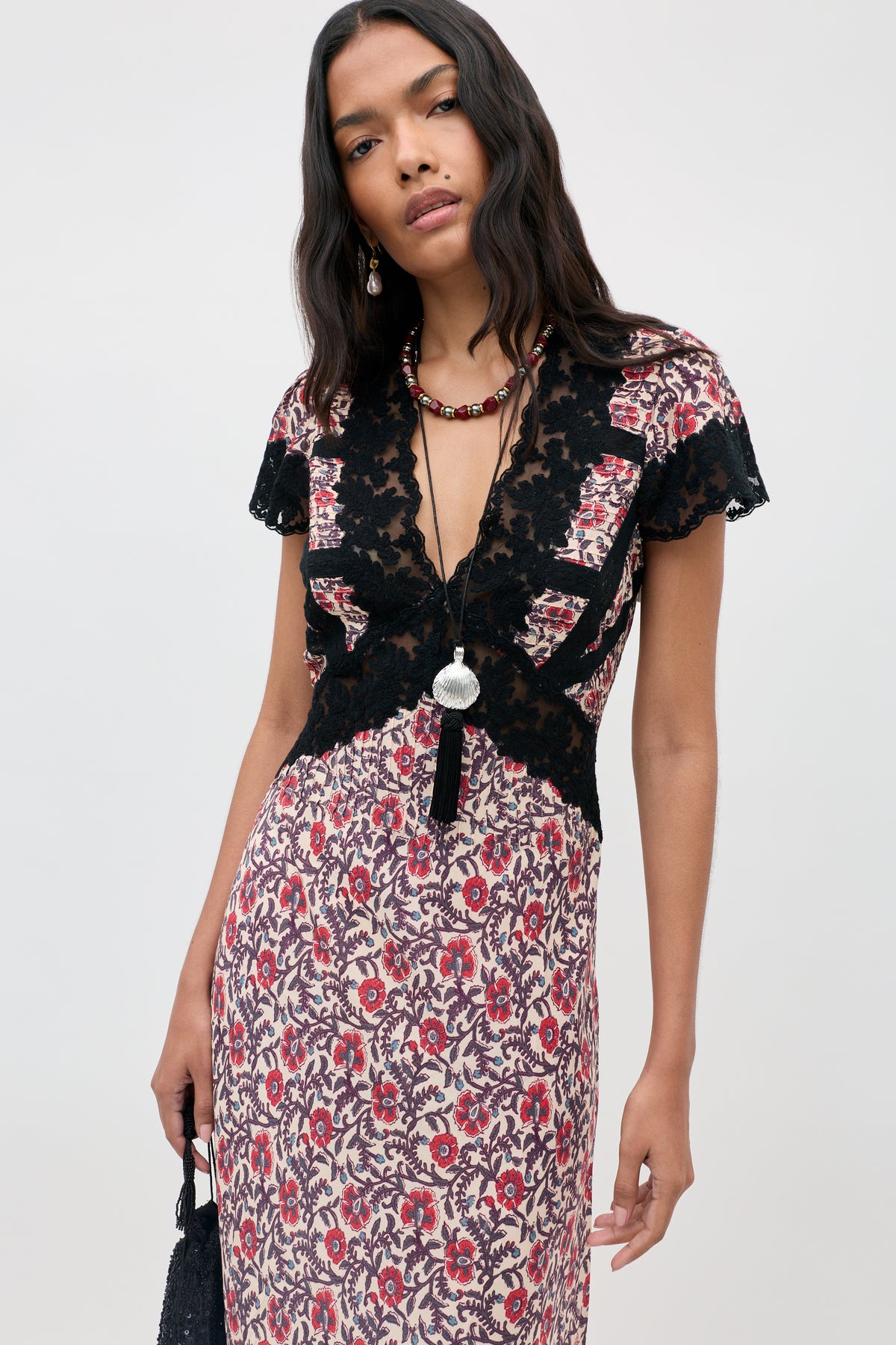 Woman wearing the Martina - Sarasa Rose Red floral dress with black lace top details, accessorized with a long necklace and earrings, holding a small black bag. She has long wavy dark hair and is standing against a plain light background.