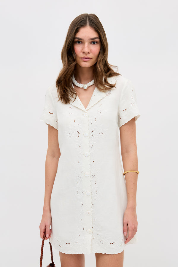 A woman with wavy brown hair wears the Zayanna - Esther Cutwork Embroidery Ivory dress, accessorized with a white necklace, gold bangle, and brown bag, standing against a plain white background.