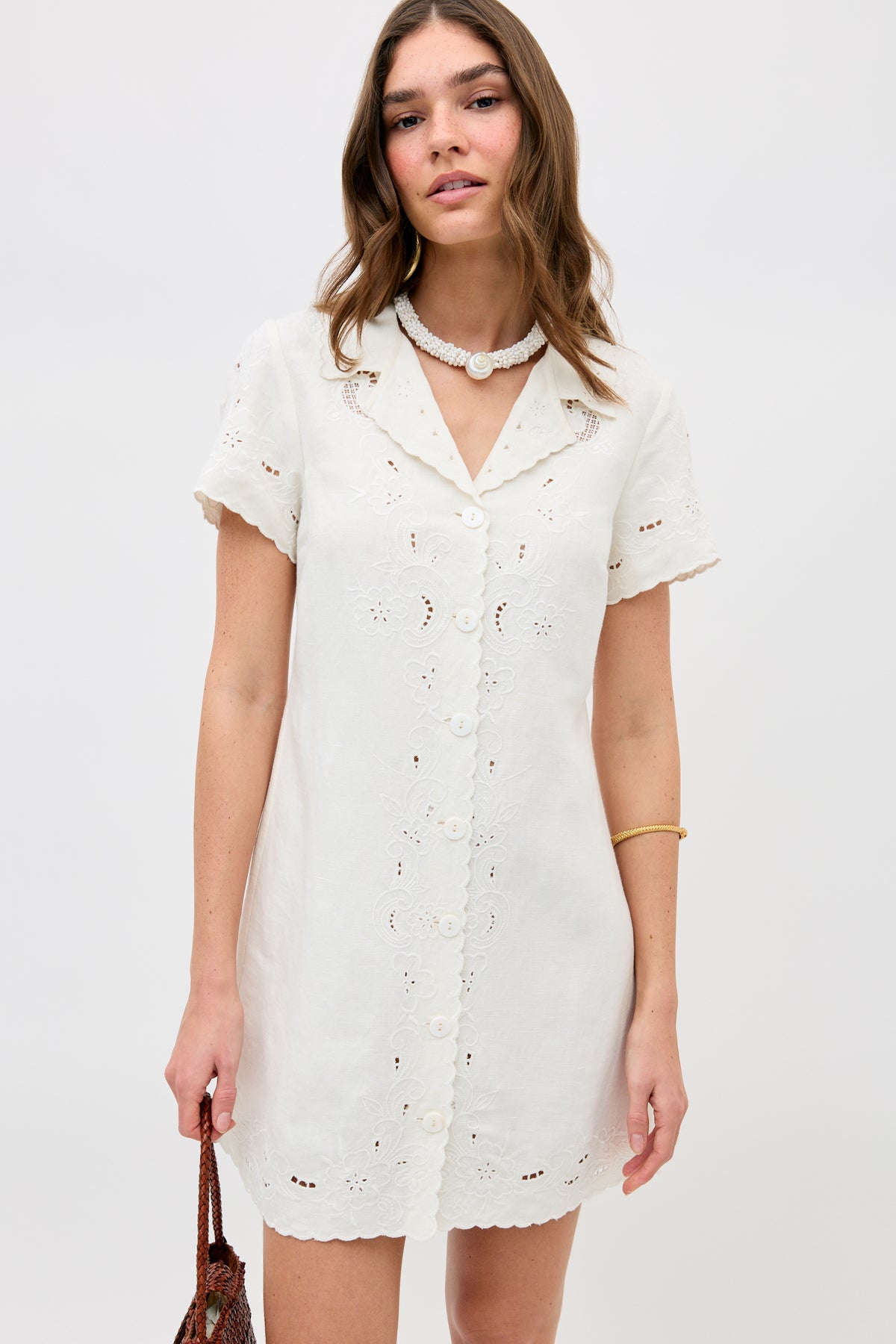 A woman stands against a plain background wearing the Zayanna Esther Cutwork Embroidery Ivory dress with short sleeves, scalloped edges, and eyelet details, styled with a brown woven bag and pearl necklace and bracelet.
