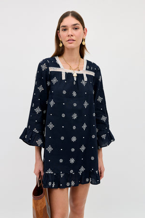 A person wears the Susana - Blossom Cutwork Embroidery Navy dress, featuring navy linen-cotton fabric adorned with white floral patterns, ruffled sleeves and hem, and delicate white lace detailing. They accessorize with a brown bag and gold jewelry while standing against a plain light background.