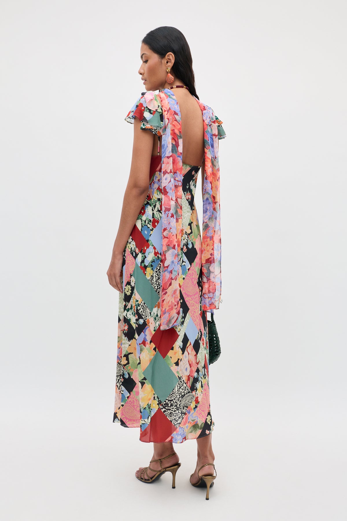 Wearing the Adele - Olivia Patchwork Multi dress, a woman stands in heels facing away. The colorful floral dress features an open back, ruffled sleeves, and draped fabric ties on the shoulders.