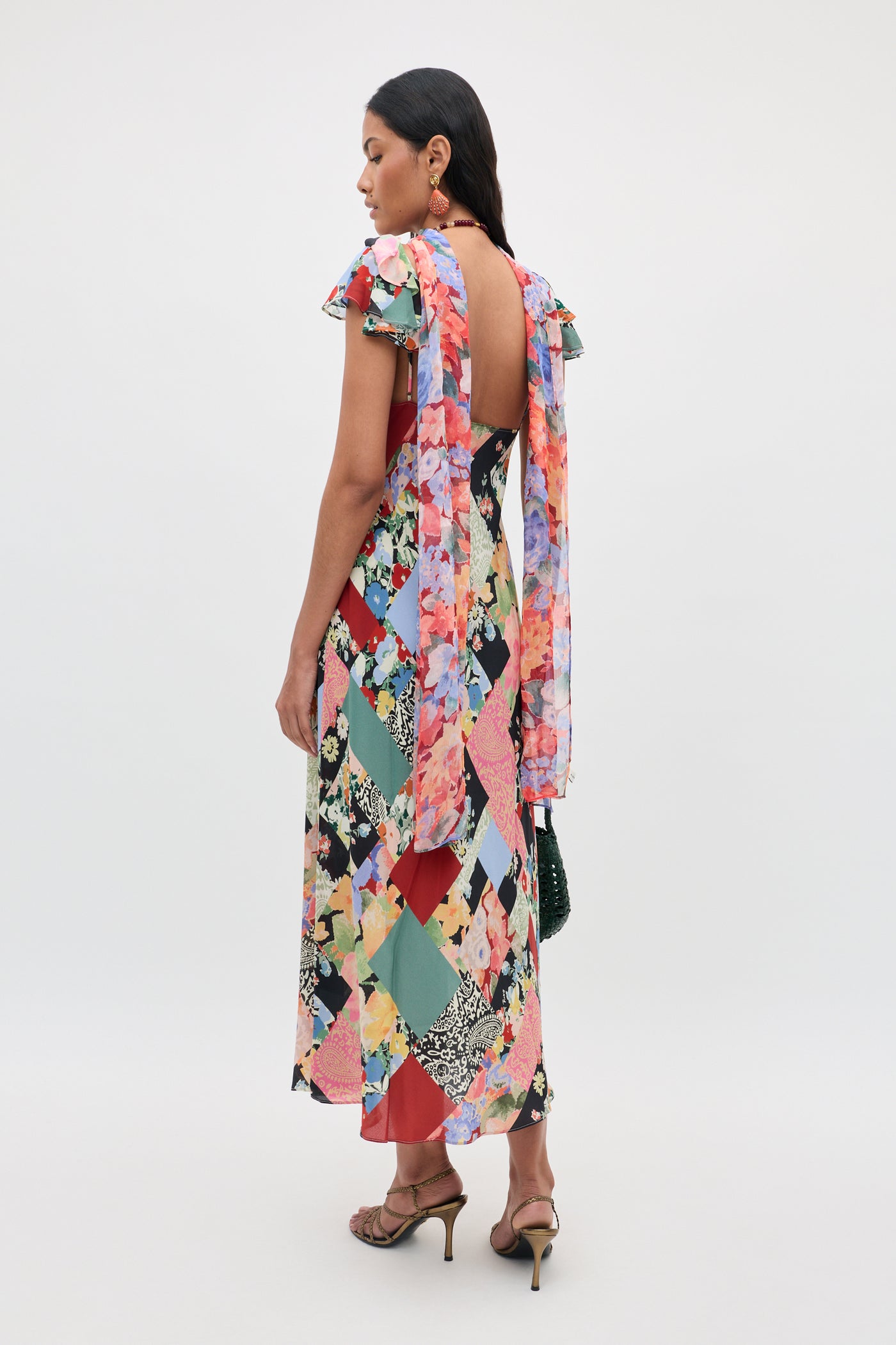 Wearing the Adele - Olivia Patchwork Multi dress, a woman stands in heels facing away. The colorful floral dress features an open back, ruffled sleeves, and draped fabric ties on the shoulders.