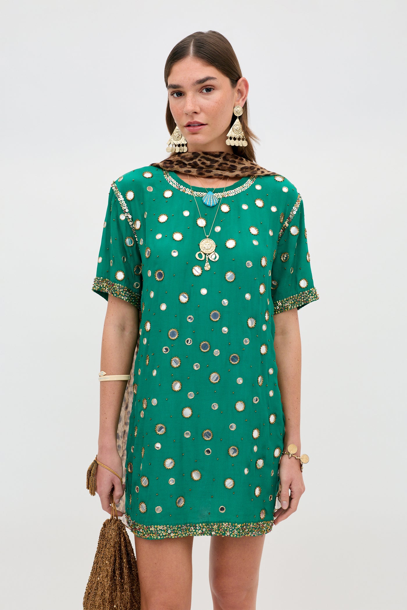 A woman stands against a plain background, wearing the Cruz - Lume Embellishment Dark Green short dress paired with gold jewelry, statement earrings, and a leopard print scarf.