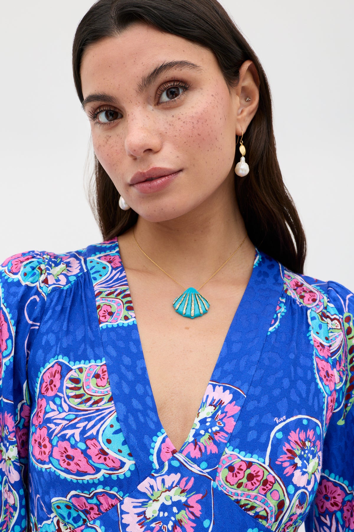 A woman with freckled skin and long dark hair wears a blue floral-patterned dress, a turquoise seashell necklace, and the elegant Rosaria - Gold drop earrings, standing against a plain light background.
