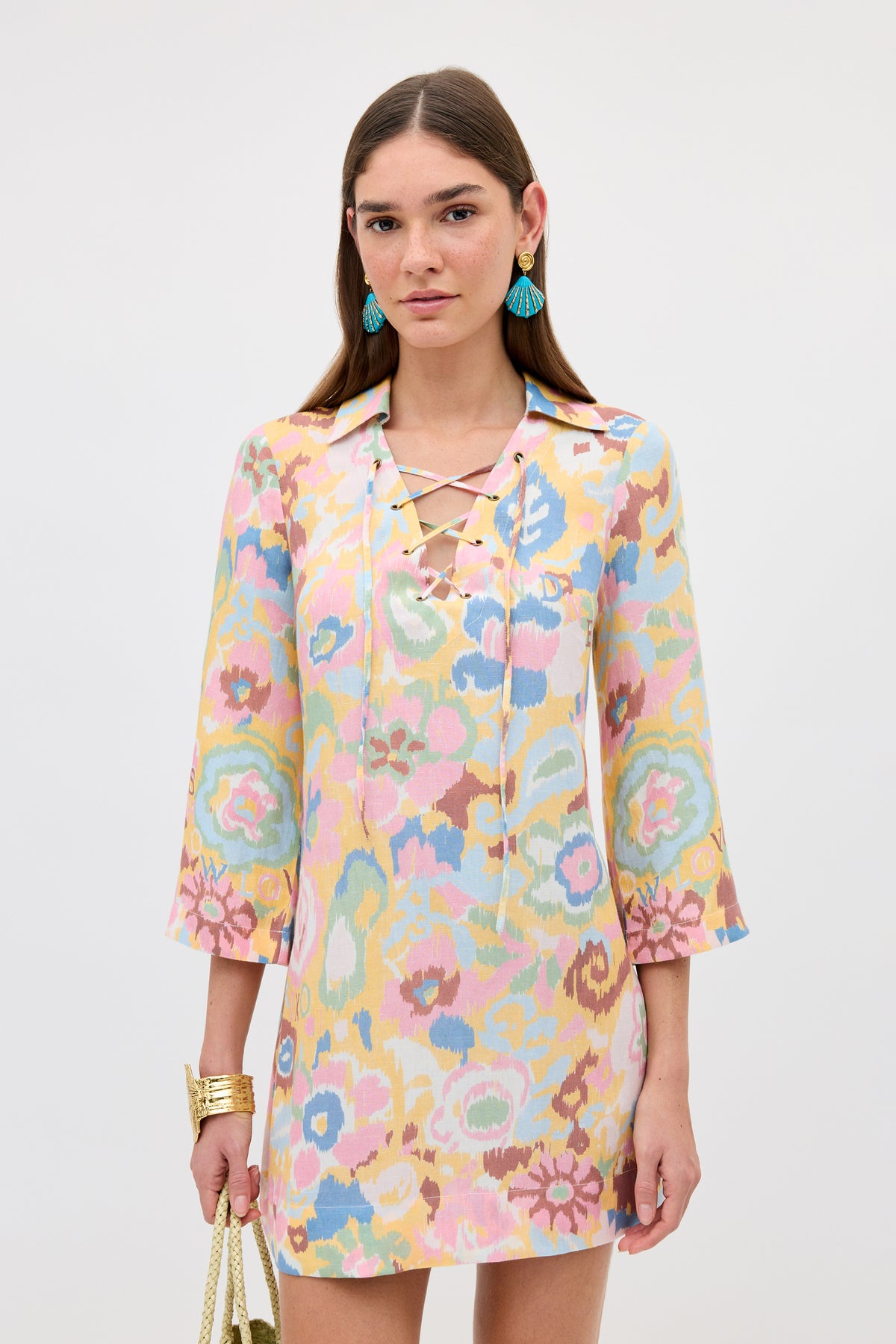 A woman with straight brown hair wears the Lucien - Ikat Bloom Yellow lace-up bohemian dress with three-quarter sleeves. She accessorizes with blue tassel earrings, gold bracelets, and a gold purse, set against a light background.