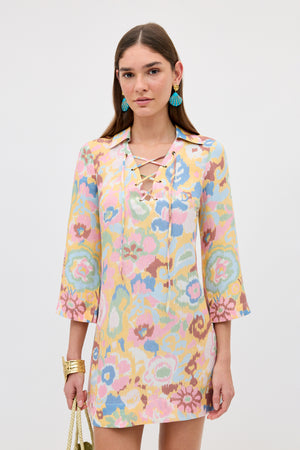 A woman with straight brown hair wears the Lucien - Ikat Bloom Yellow lace-up bohemian dress with three-quarter sleeves. She accessorizes with blue tassel earrings, gold bracelets, and a gold purse, set against a light background.