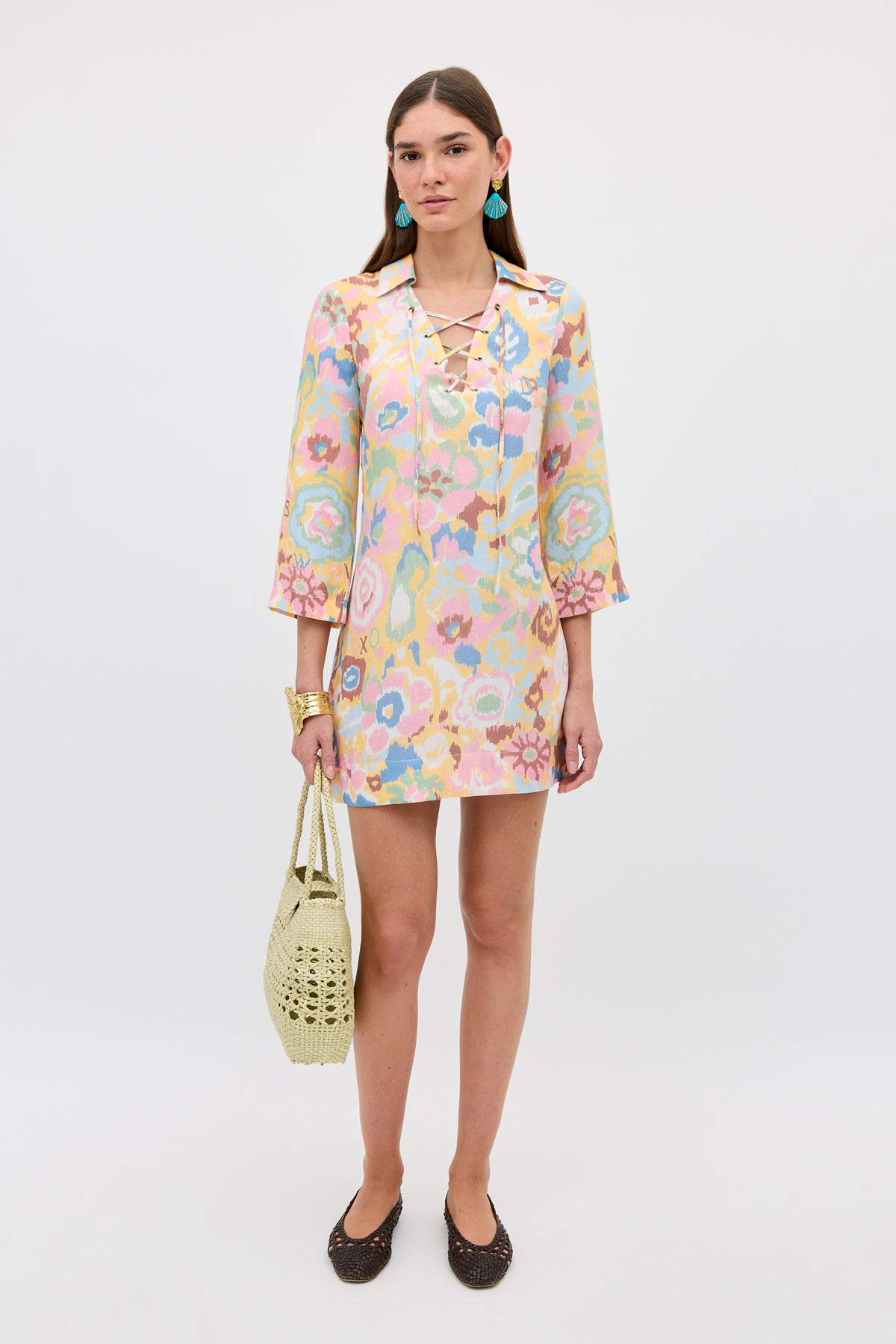 A woman stands against a plain background wearing the Lucien - Ikat Bloom Yellow linen mini dress with lace-up front, brown flats, turquoise earrings, gold bracelets, and holding a woven beige bag.